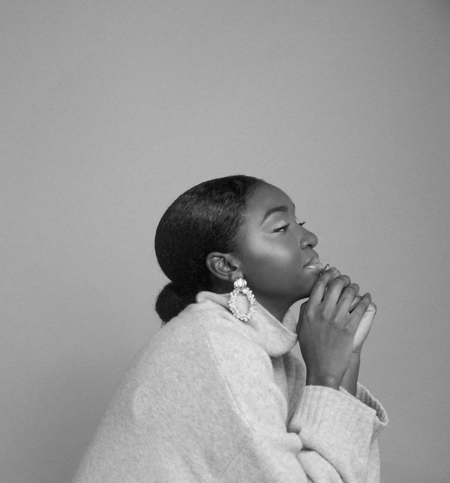 Black and white photo of a woman with her hair neatly styled, wearing a light-colored sweater, with her eyes closed and hands clasped near her face, wearing large circular earrings.