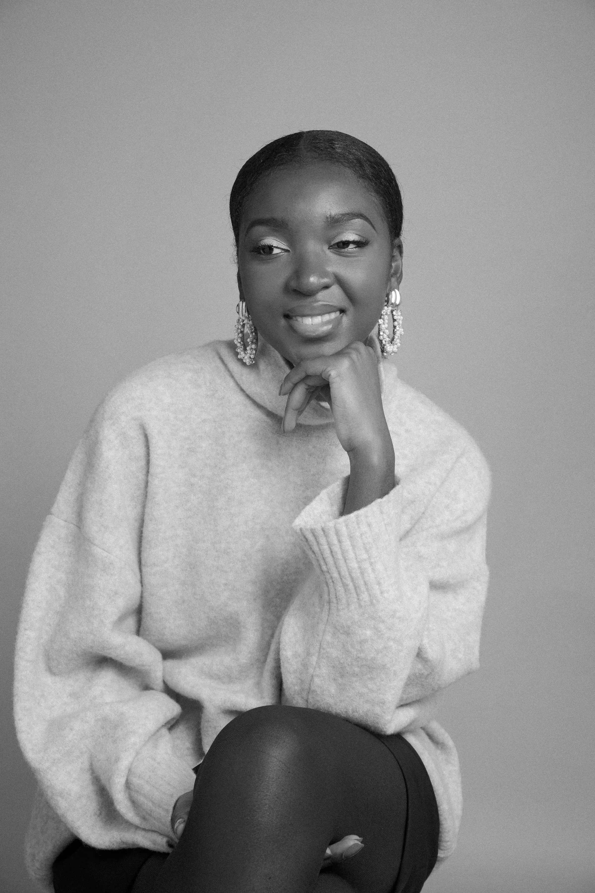 Black and white portrait of a woman with short hair, wearing a light-colored sweater, earrings, and sitting with her hand under her chin, holding a black hat in her lap, against a plain background.