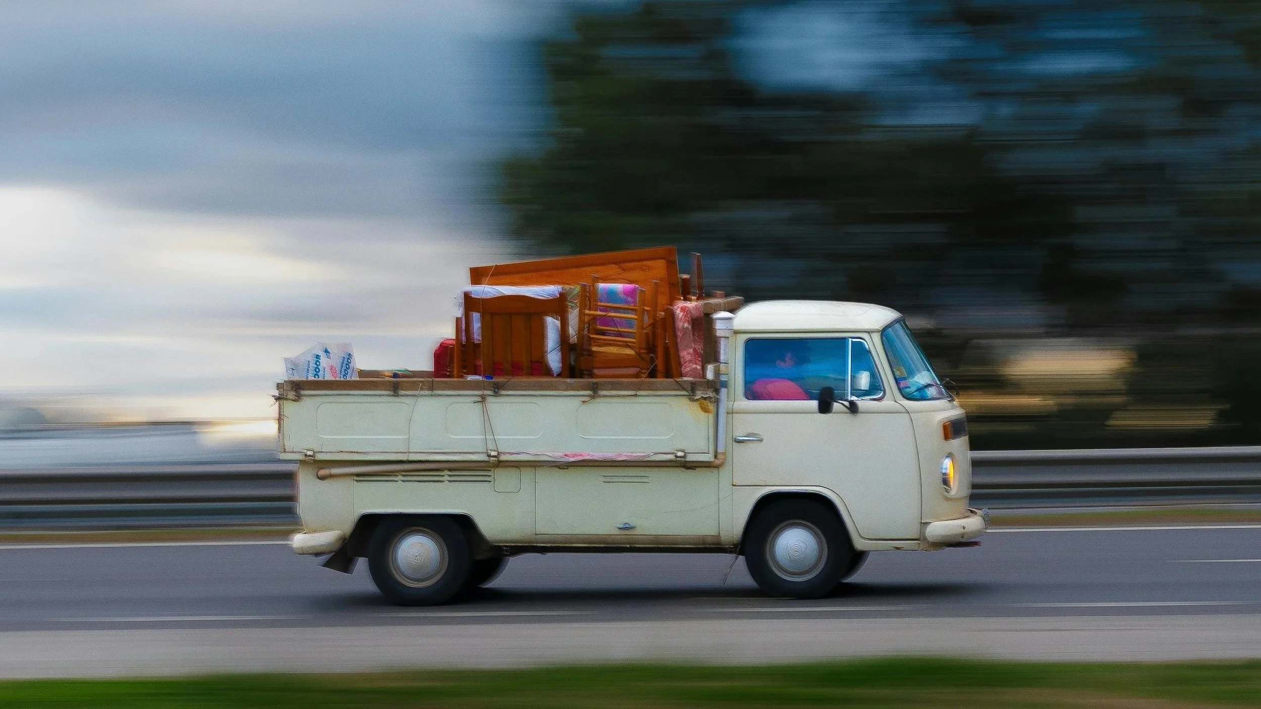 Ein weißer Kleintransporter fährt auf der Autobahn, beladen mit Stühlen, einem Holzregal und Verpackungskartons, während der Himmel bewölkt ist.
