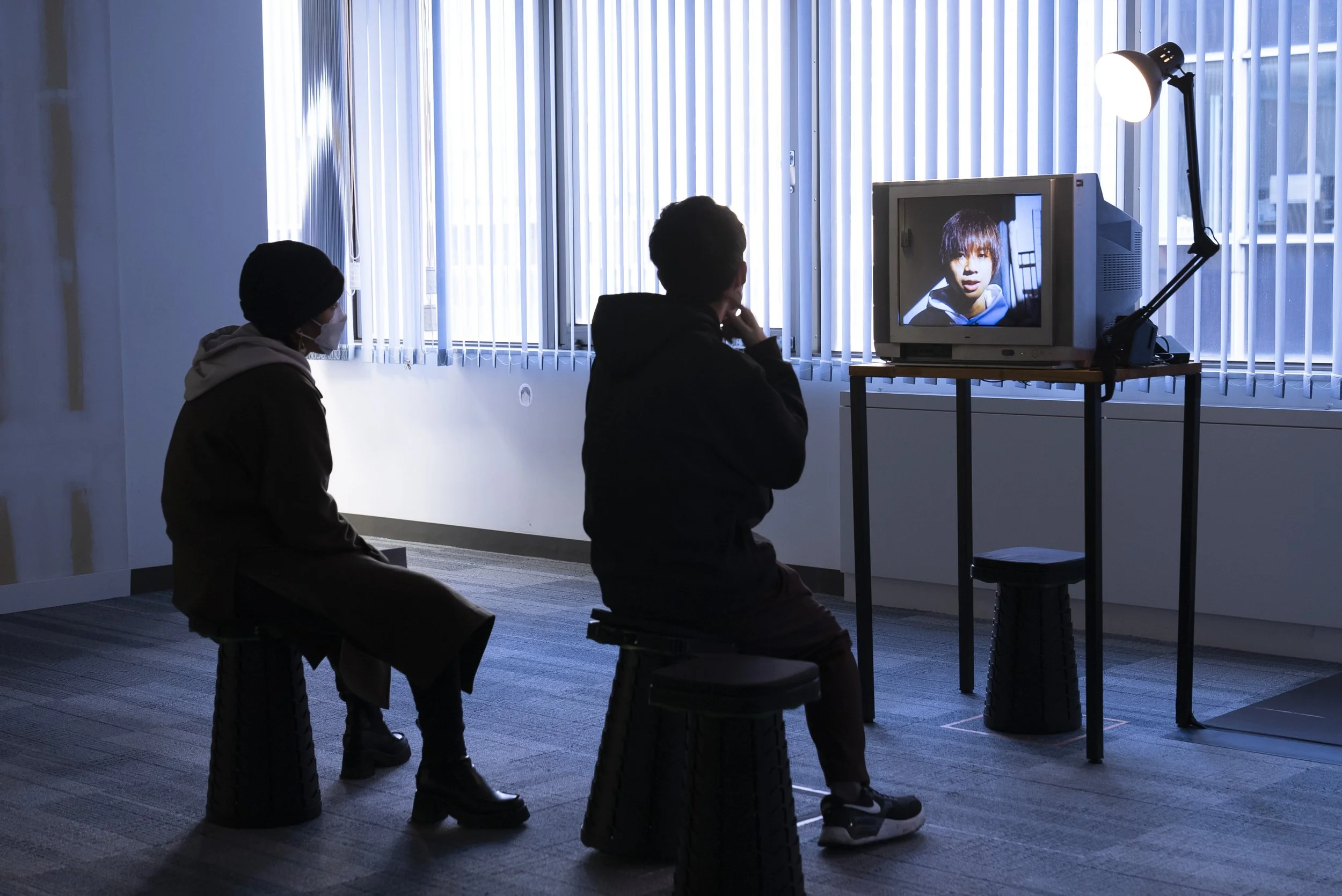 二人の人がコンテナのテレビ画面を見ている室内のシーン、窓からの自然光と人工照明が映る