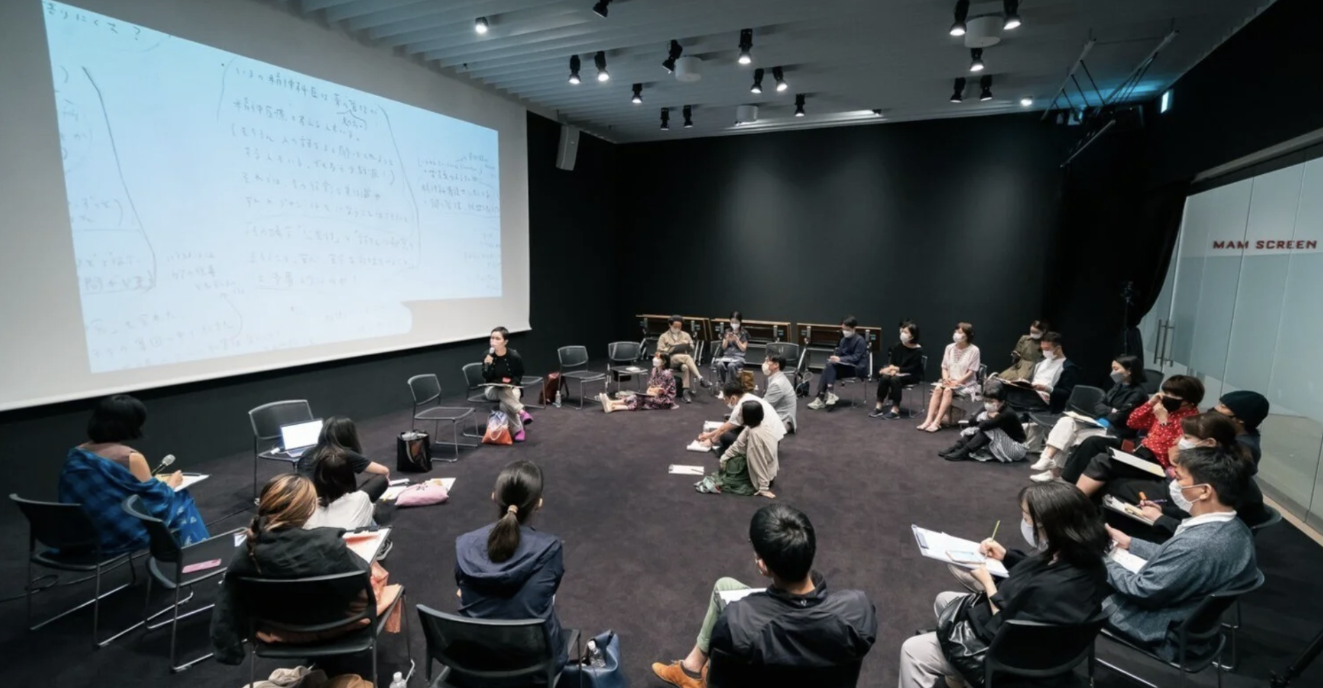 黒い壁の前に円形に配席された多人数の参加者が座っている会議やワークショップの風景。壁側に大きなスクリーンに書かれたホワイトボードや資料が映し出されている。多くの参加者がフェイスマスクを着用し、メモを取ったり見たりしている。