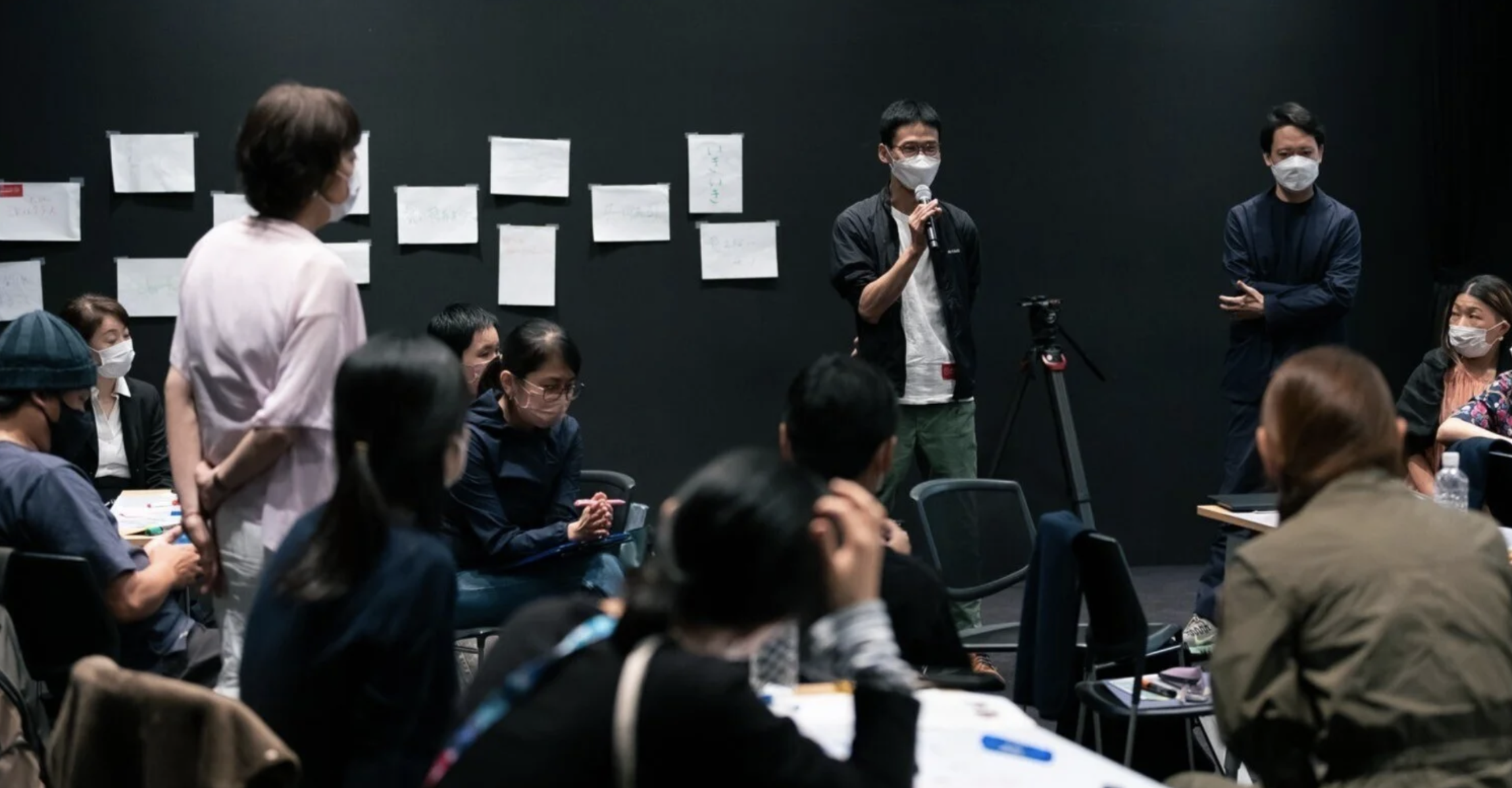 会議やプレゼンテーションを行う人々の集まり。黒い背景にホワイトボードや紙が貼られ、多くの参加者がマスクを着用して座っている。