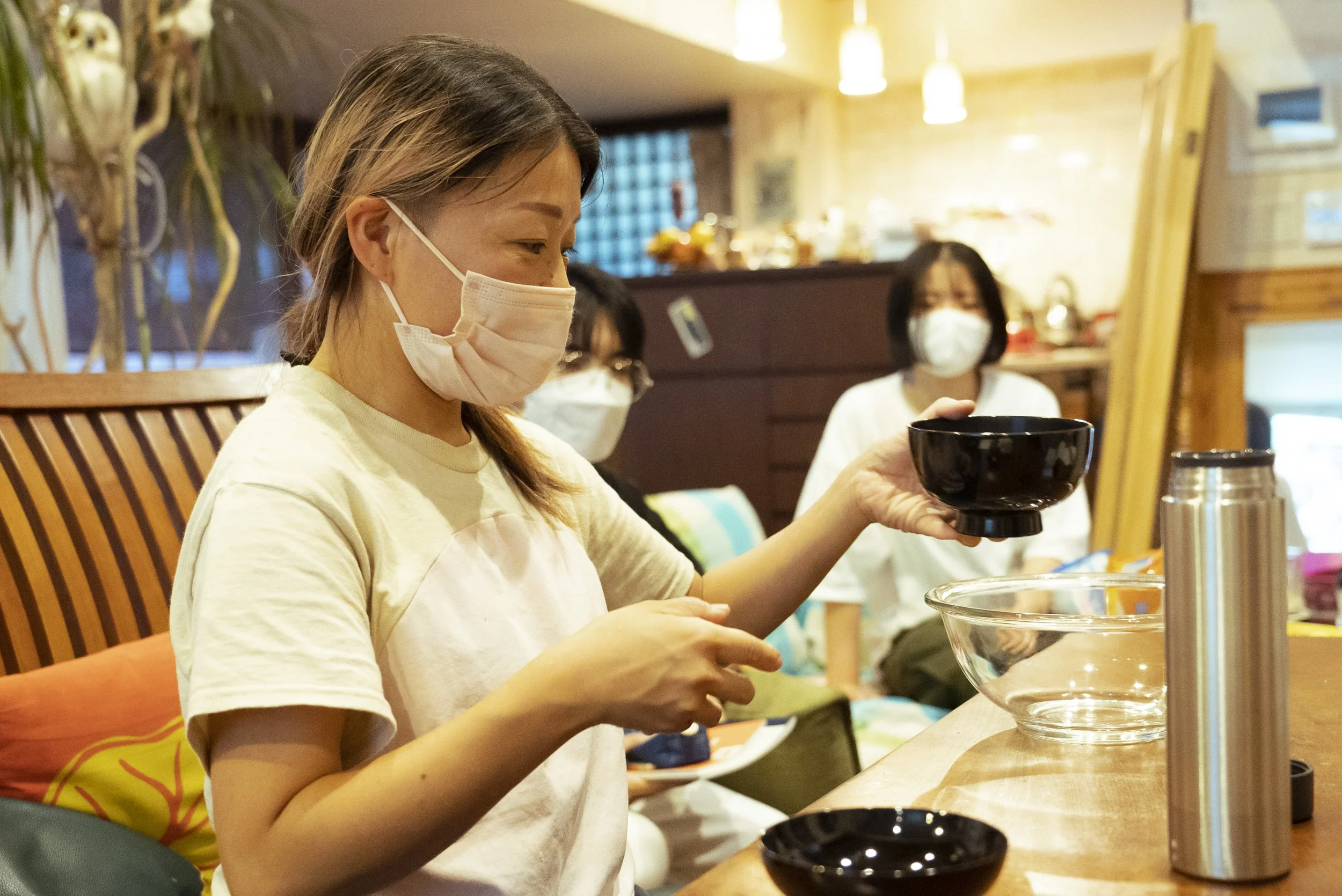 複数の女性がマスクを着用し、料理や食事の準備をしている室内のキッチンやダイニングエリアの光景