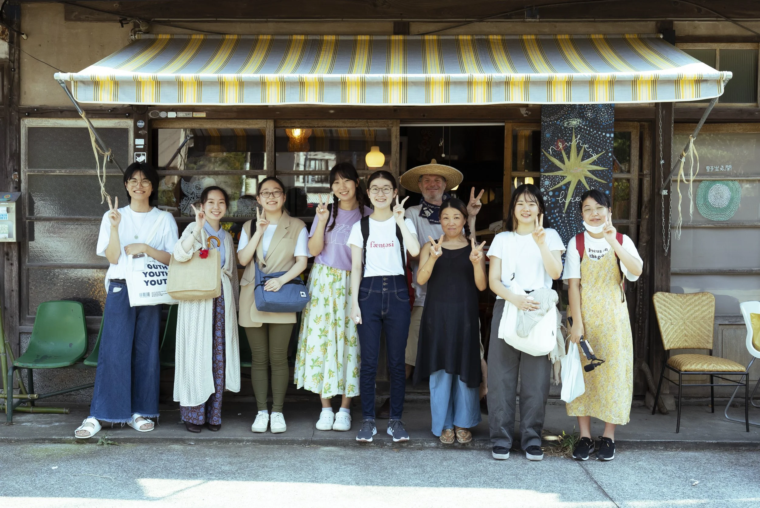 多人数の女性と男性が笑顔でポーズを取りながら外で写真を撮影している。背景は木造の建物とカラフルな装飾が施された壁。