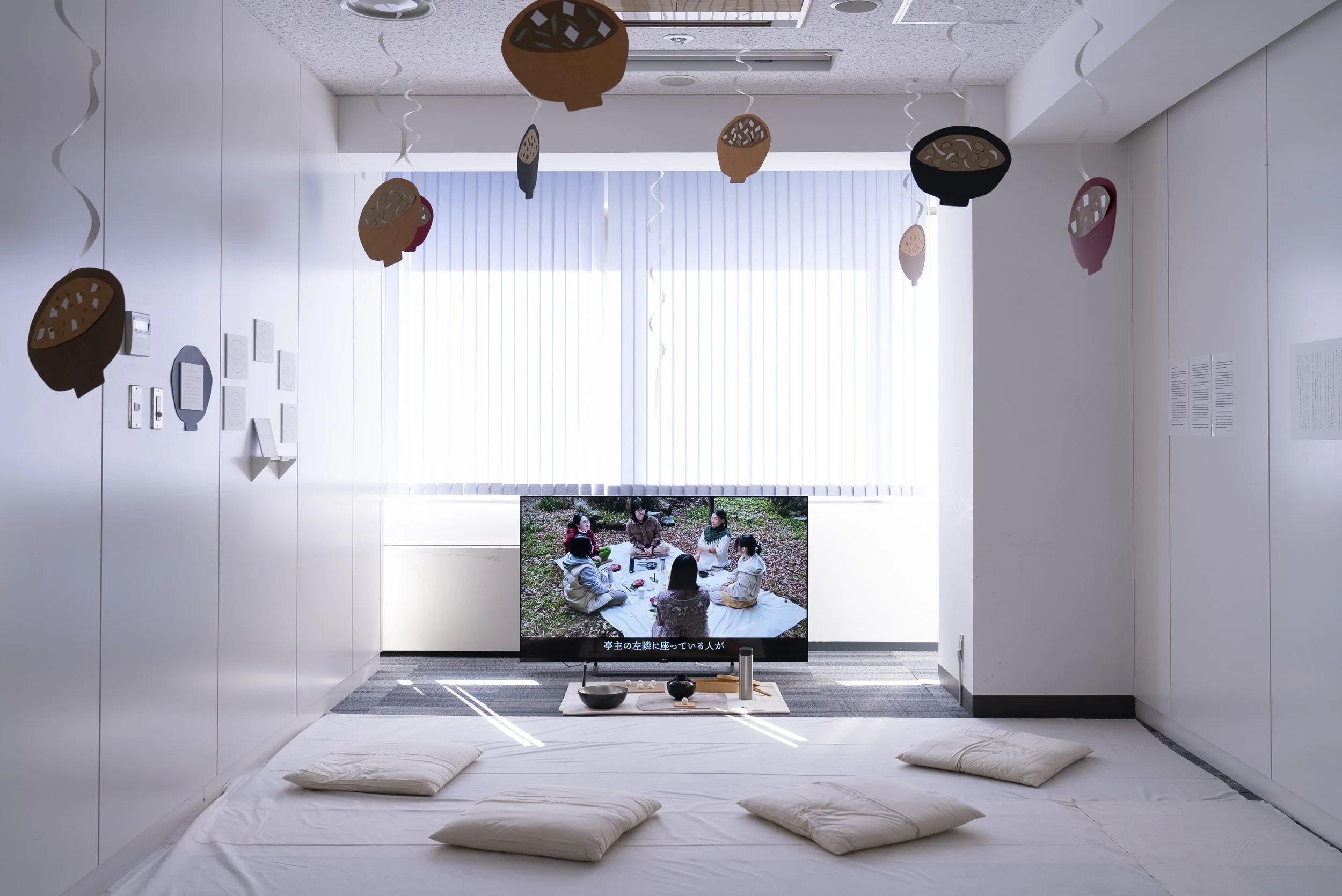 白い部屋に布団と枕が敷かれた床があり、テレビ画面には屋外で座る人々の映像が映っている。天井には吊るされた飾りがいくつかあり、窓には縦長のブラインドがある。