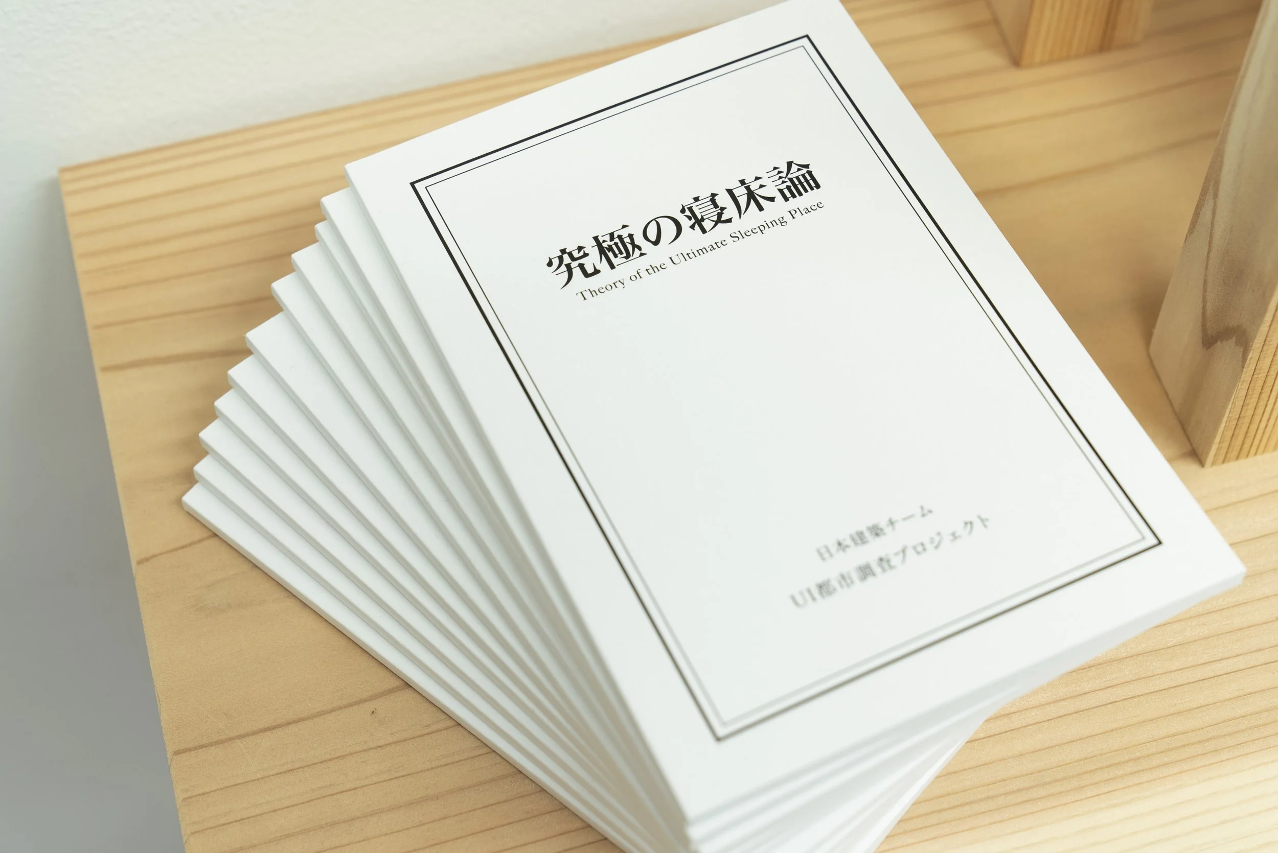 白い表紙の本が積まれた本棚の上に置かれている。表紙には日本語と英語で「究極の寝床論」と記載されている。