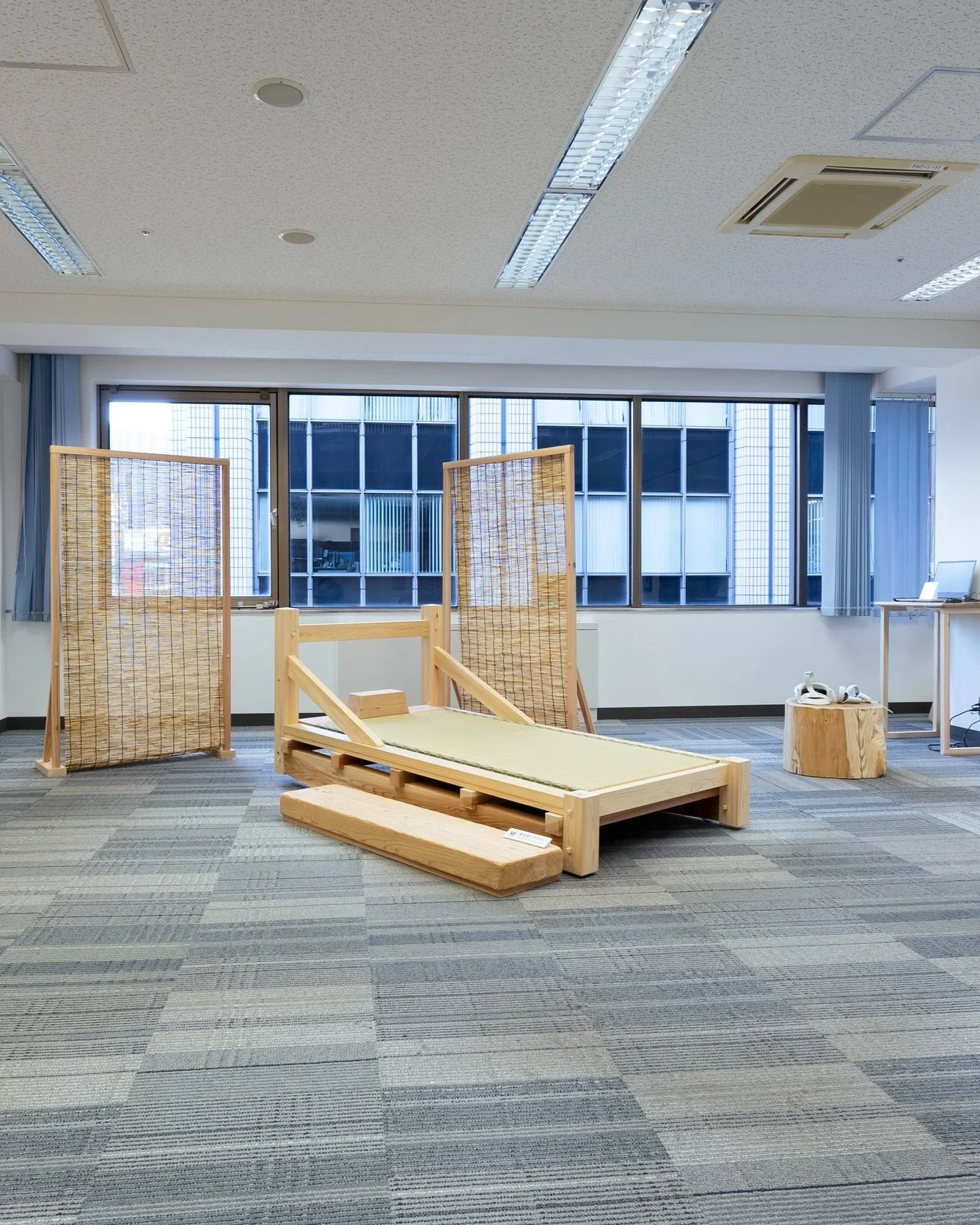 室内に木製の段差と仕切りが置かれたスペース。背景には窓とビルの外観が見える。