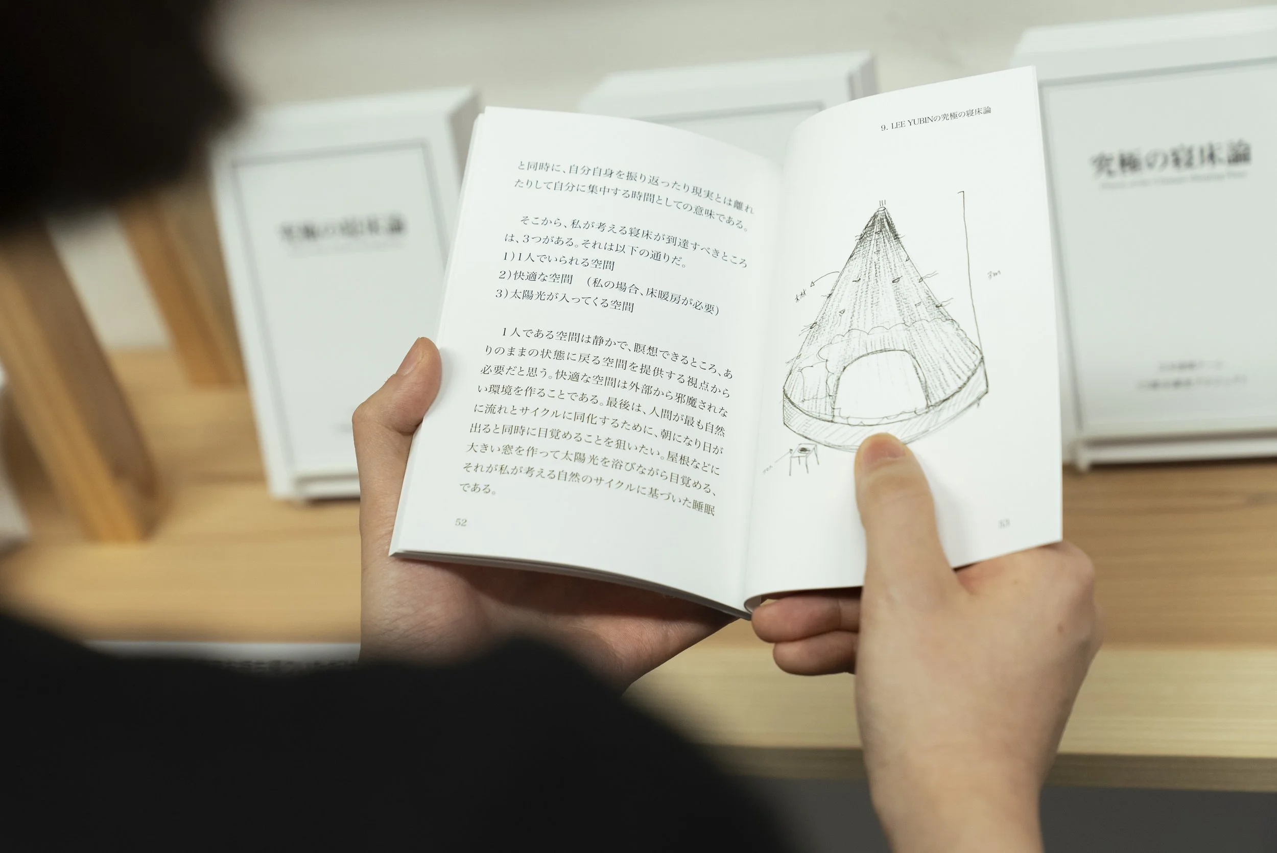 人が本を読んでいる。ページにはドーム型の構造のスケッチと日本語の文章が書かれている。背景には他の同じ本が積まれている。