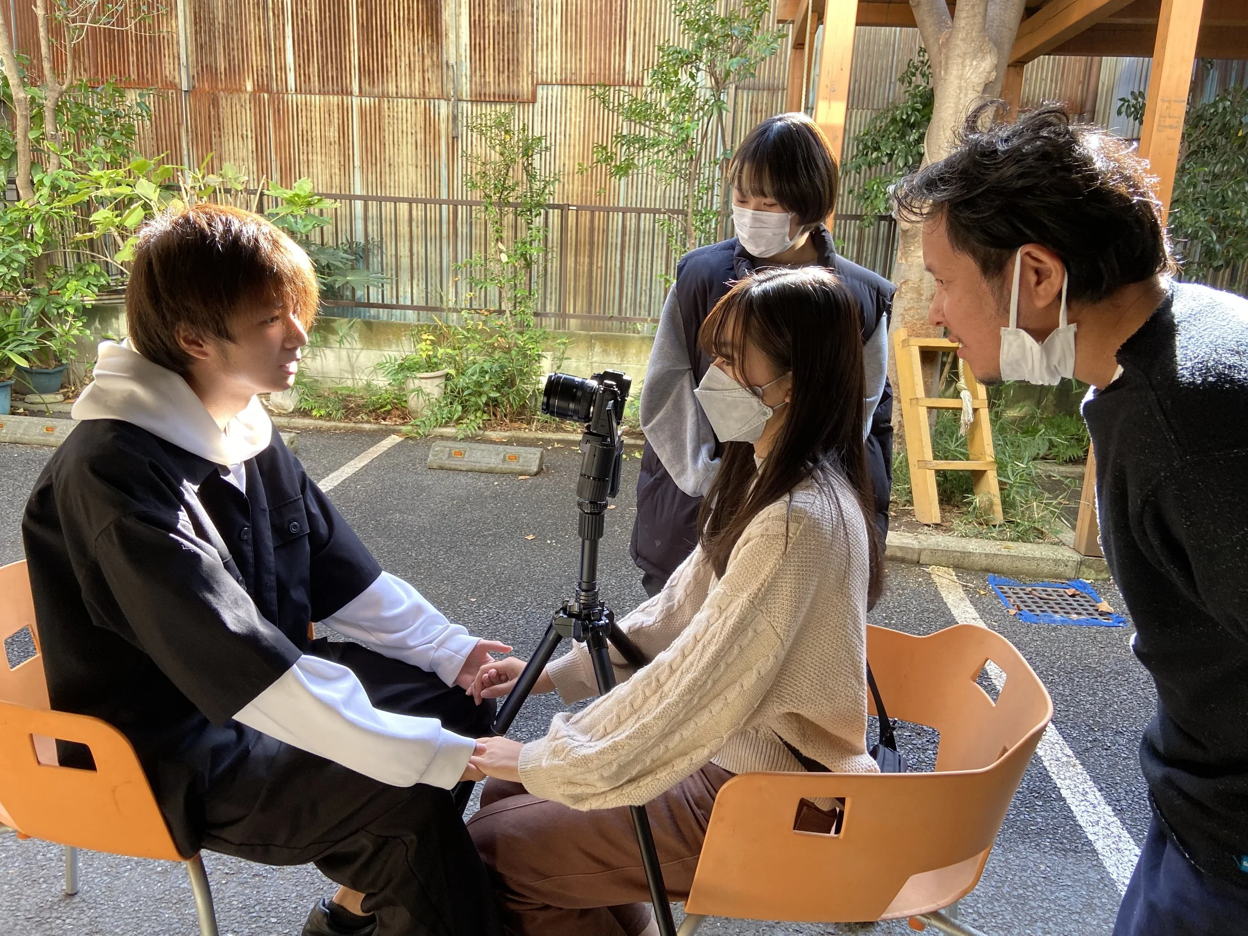 屋外で映像撮影をしているシーン。女性二人が椅子に座り、一本のカメラを持ちながら話している。二人ともマスクをしている。髪や背景の木々が見える様子。