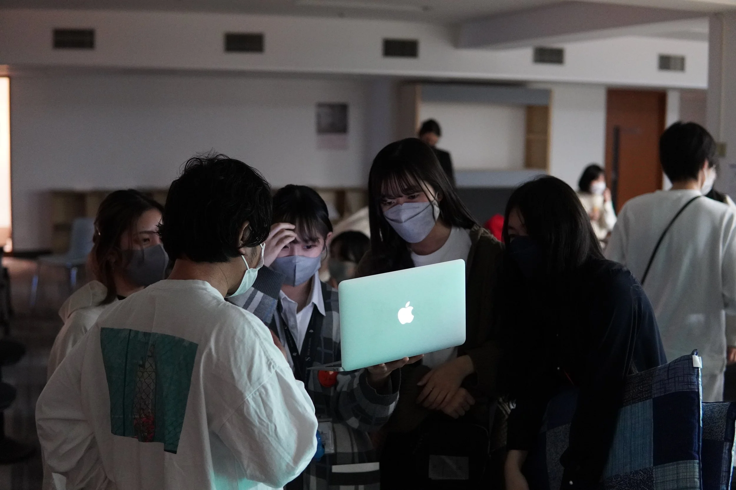 マスクをした若い女性と男女がAppleのロゴが表示されたノートパソコンの前で話し合っているグループの様子