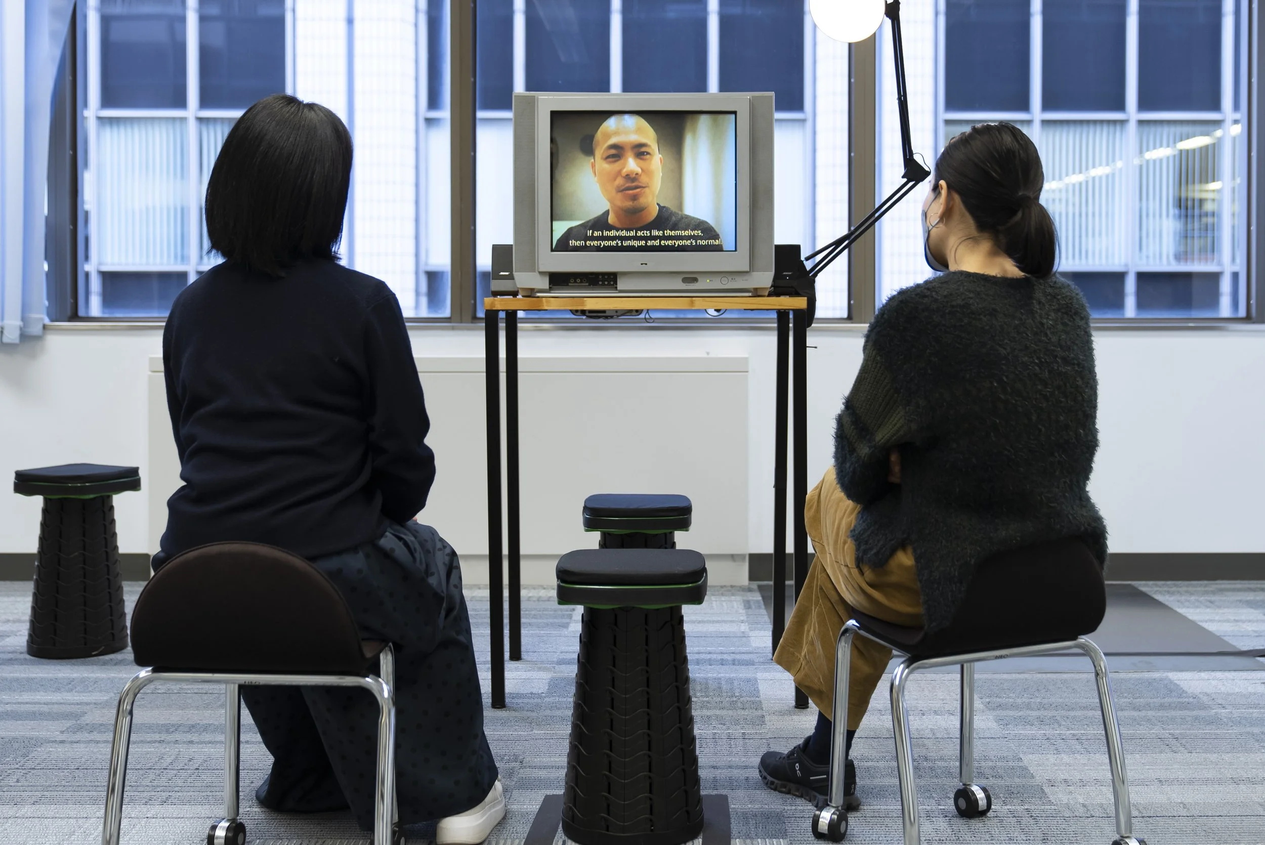 2人の女性が座って見ているテレビ画面に、男性の顔と字幕が映っている室内の様子