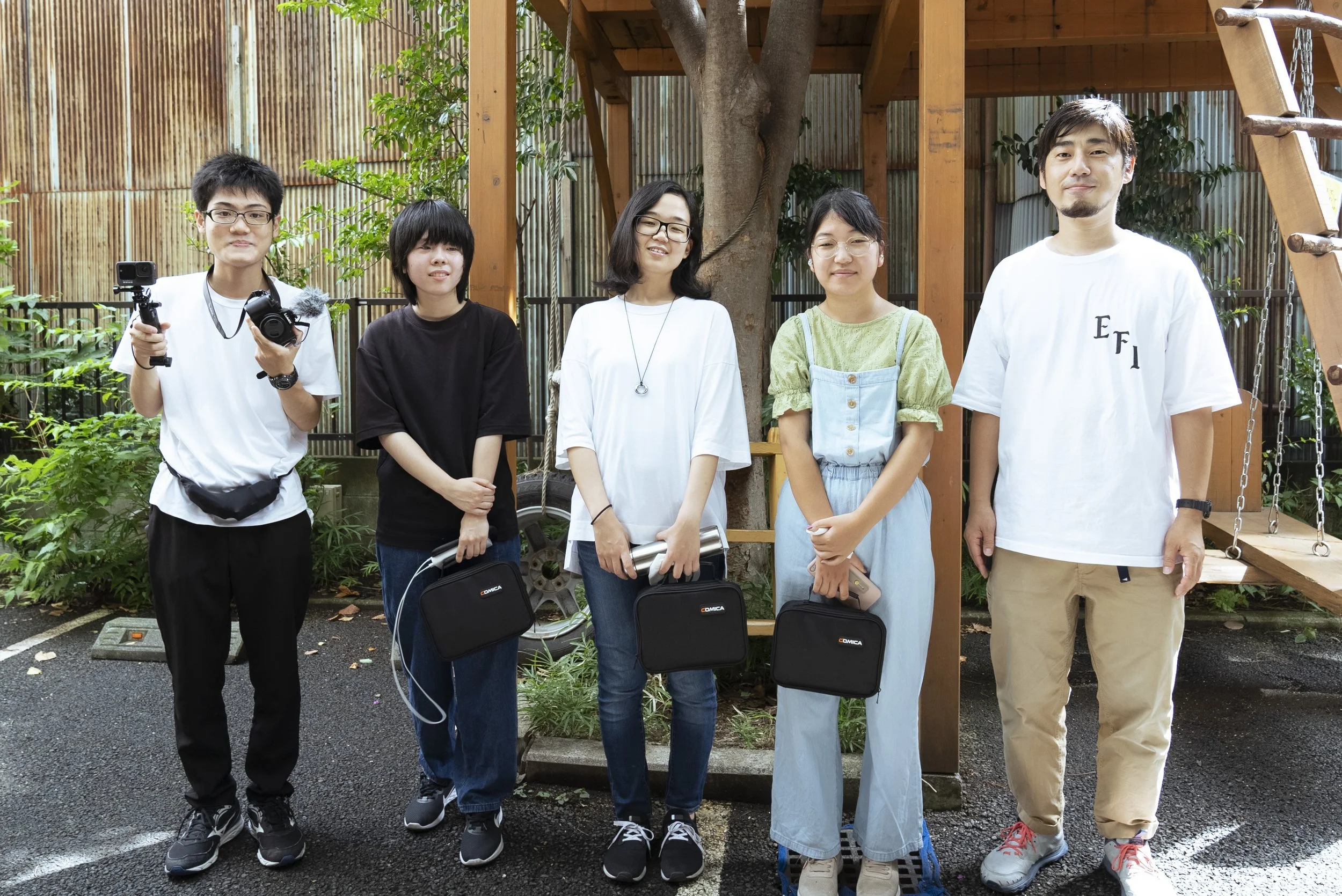 屋外の庭で、五人の若者がカメラと映像機器を持ち、並んで立っている。背景には木と竹のフェンス、遊具のブランコと木製の遊具が見える。皆笑顔で casualな服装をしている。
