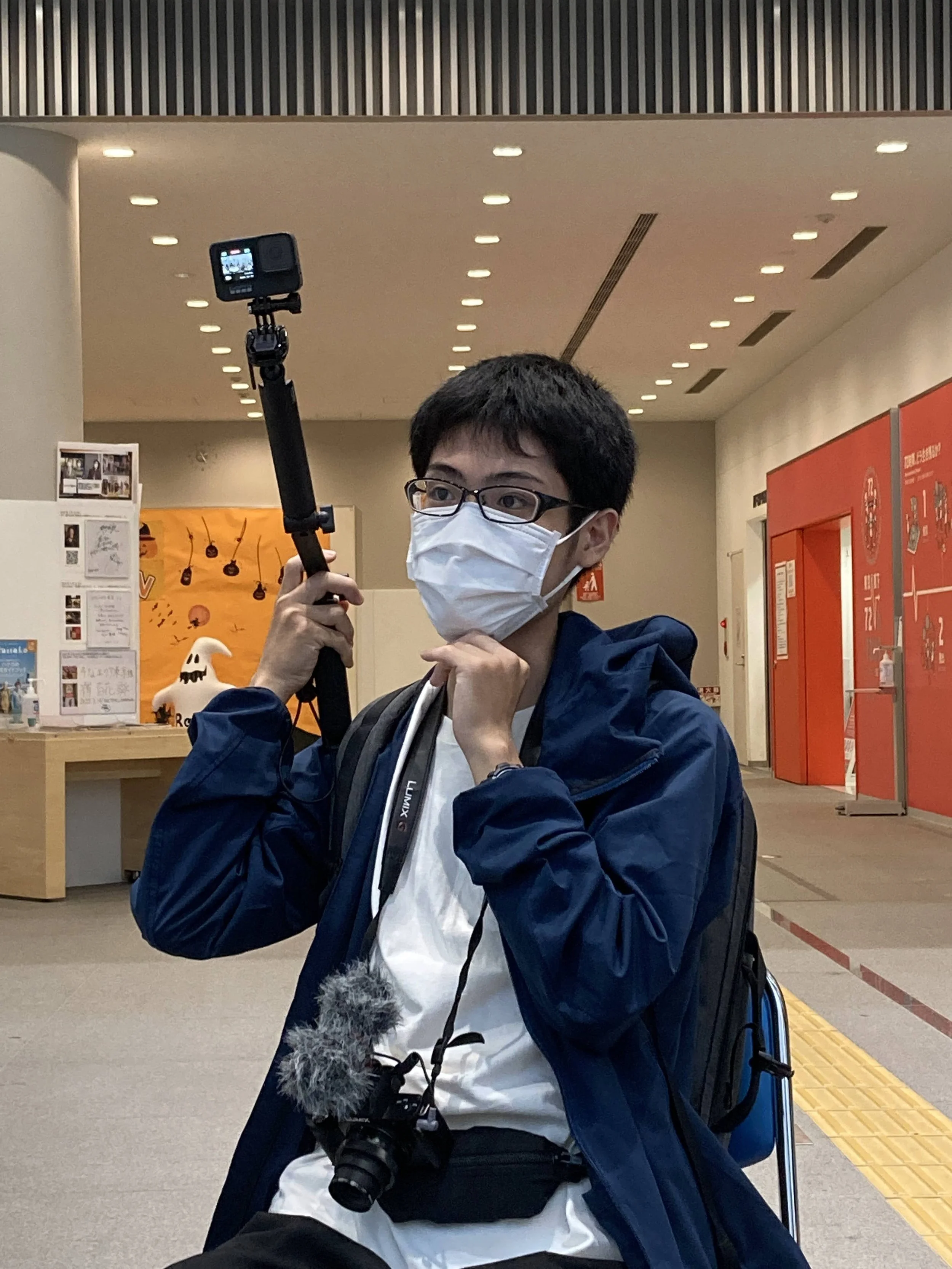 マスクをしている男性が、自撮り棒を使って写真を撮っている。背景は室内で、赤い看板や展示物が見える。