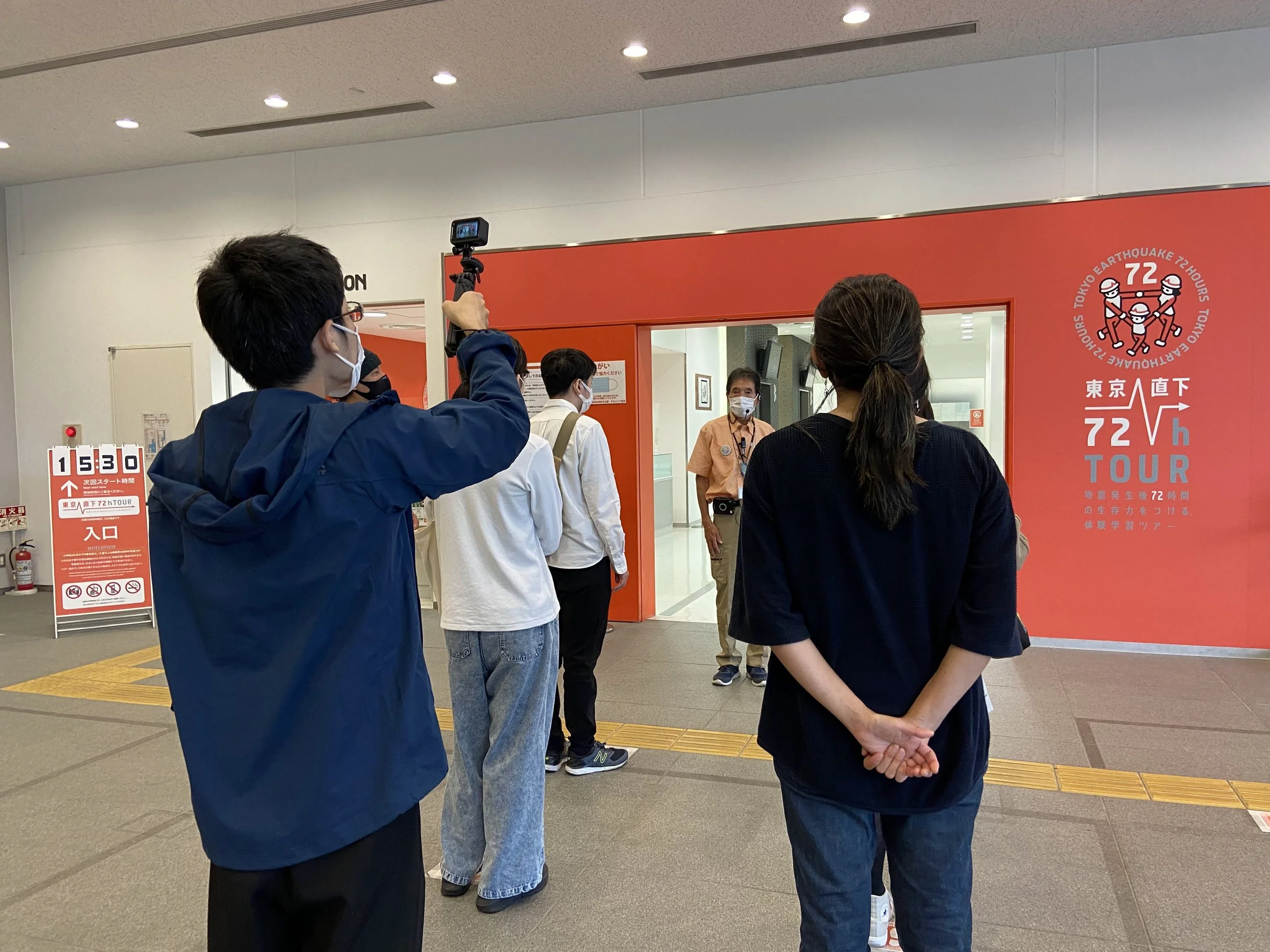 東京直下72時間耐震体験学習ツアーの案内マークと入口付近の人々の写真。