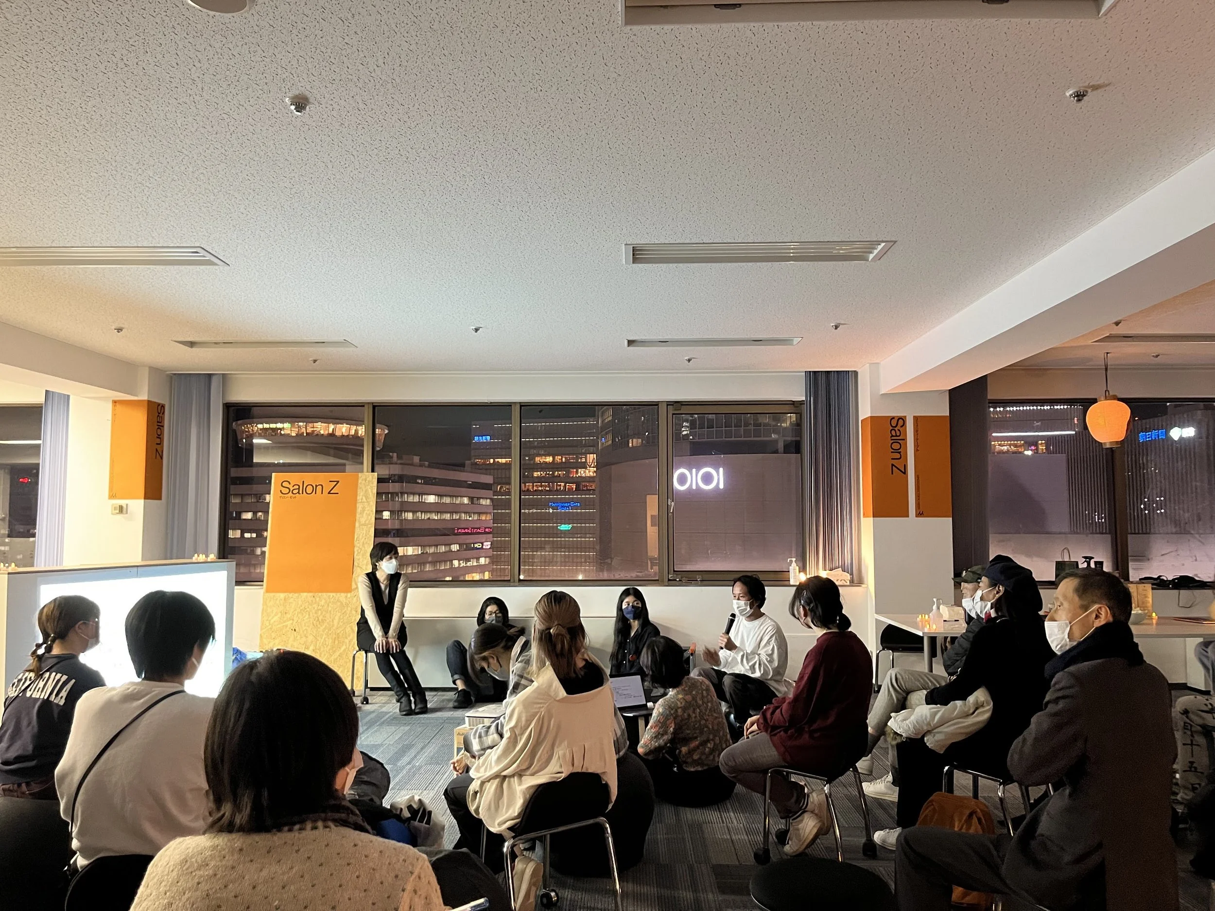 室内で行われているトークイベントの様子。参加者はマスクを着用しており、座って話を聞いている。背景には大きな窓と夜景が見える。