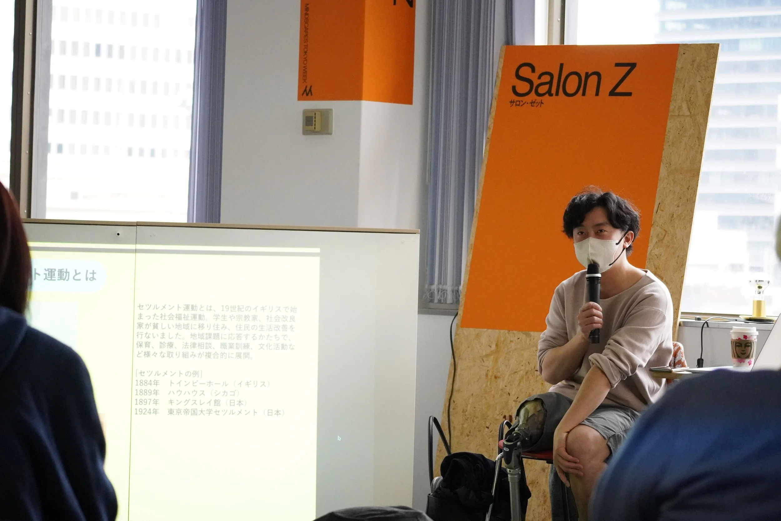 室内の講義中の若い男性講師がマスクをして、マイクを手に持って話している。背景にオレンジ色の掲示板に「Salon Z」と書かれている。白いスクリーンに日本語のプレゼンテーション資料が映し出されている。