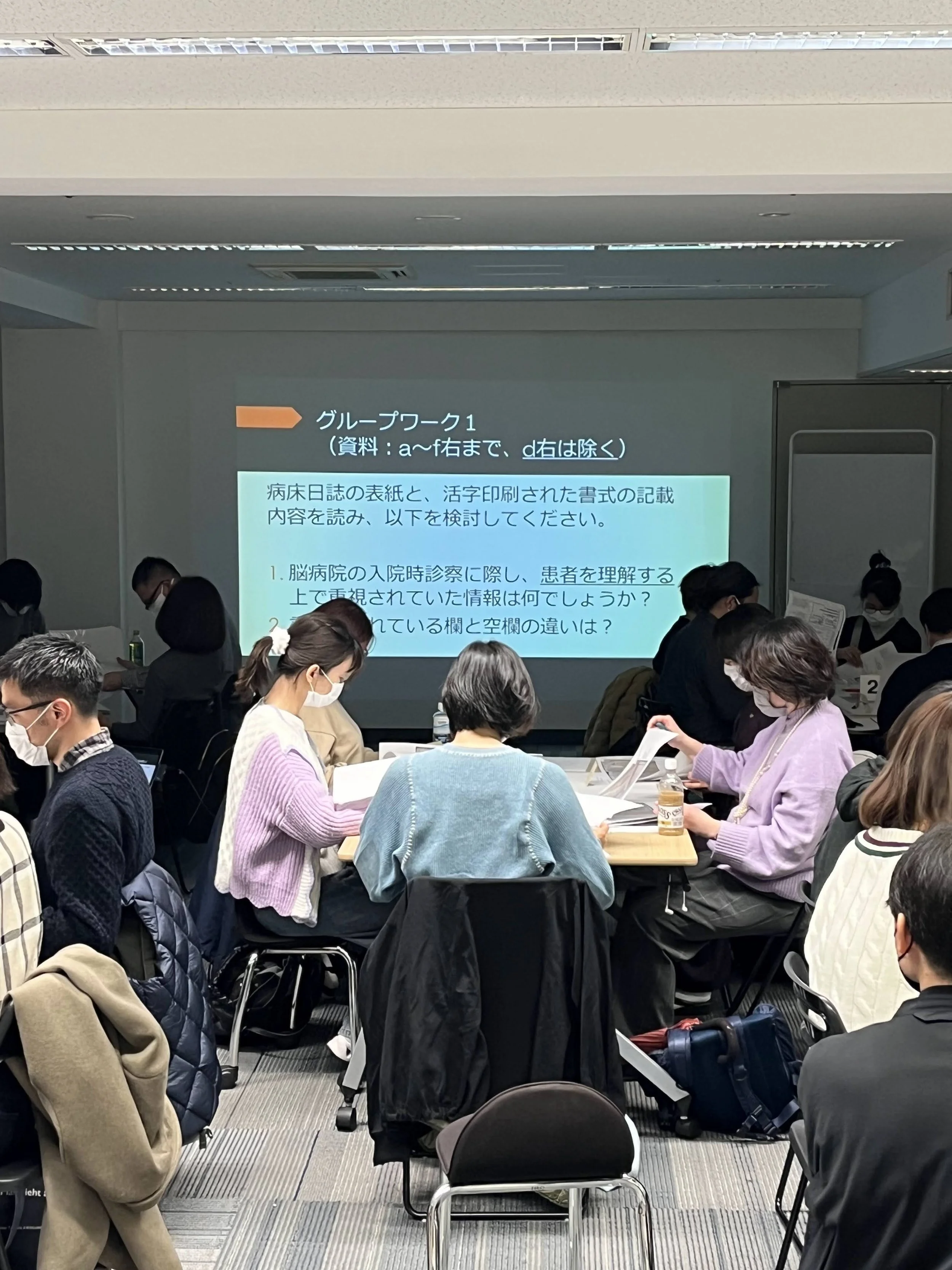 会議室で参加者たちが資料を読みながら座っている。スクリーンに資料の内容が映し出されている。