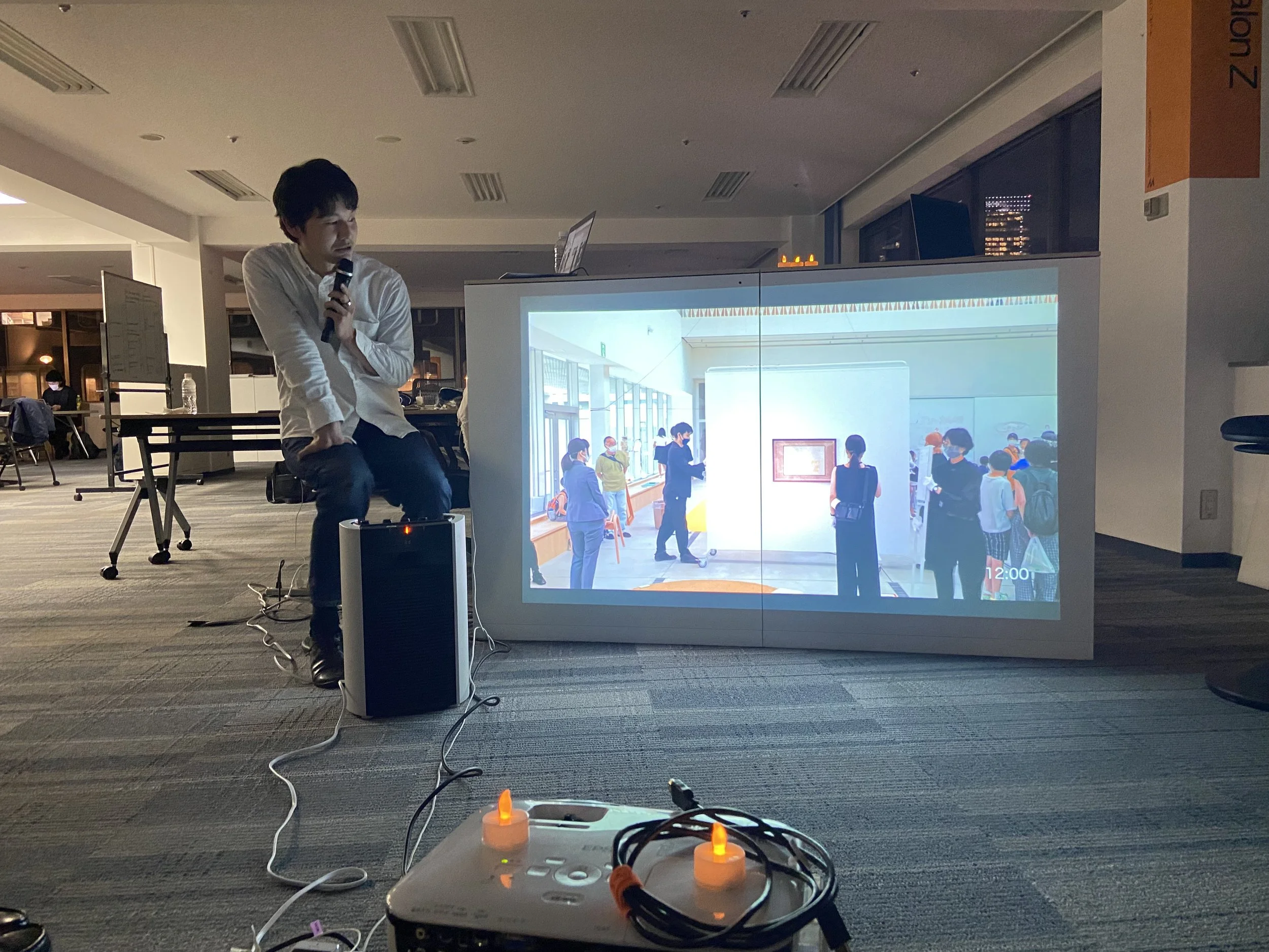 スライドの前でマイクを持って話す男性と、スクリーンに映し出された展示会場の画像