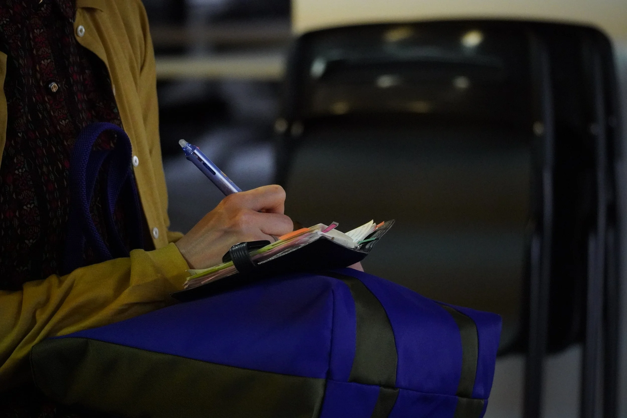 バッグを持った女性がメモ帳に書き込みながら座っている。背景に椅子が見える。