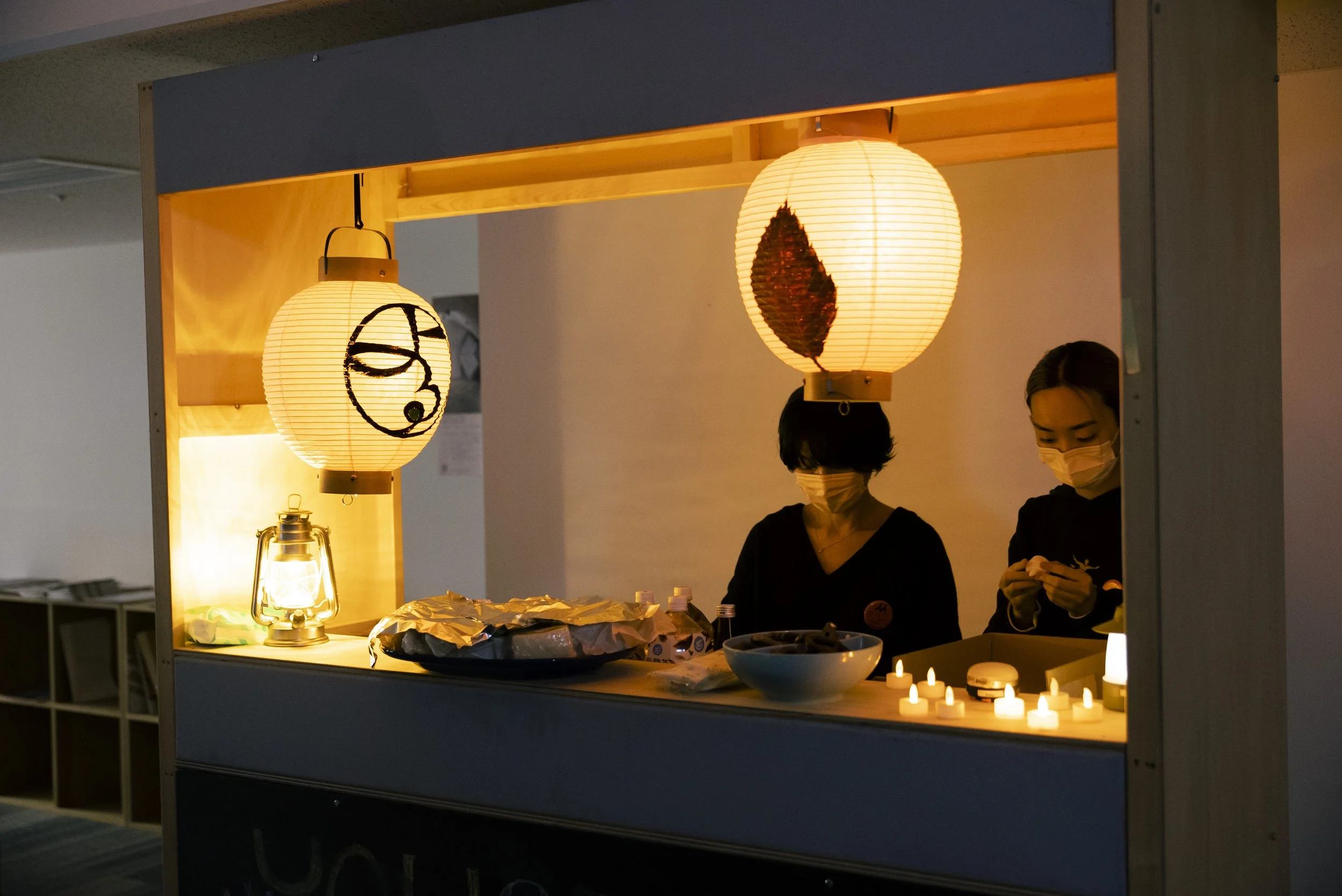 二人の女性が屋台のような小さな照明付きのカウンターで作業している。ふたりともマスクを着用し、和風の提灯が吊るされている。