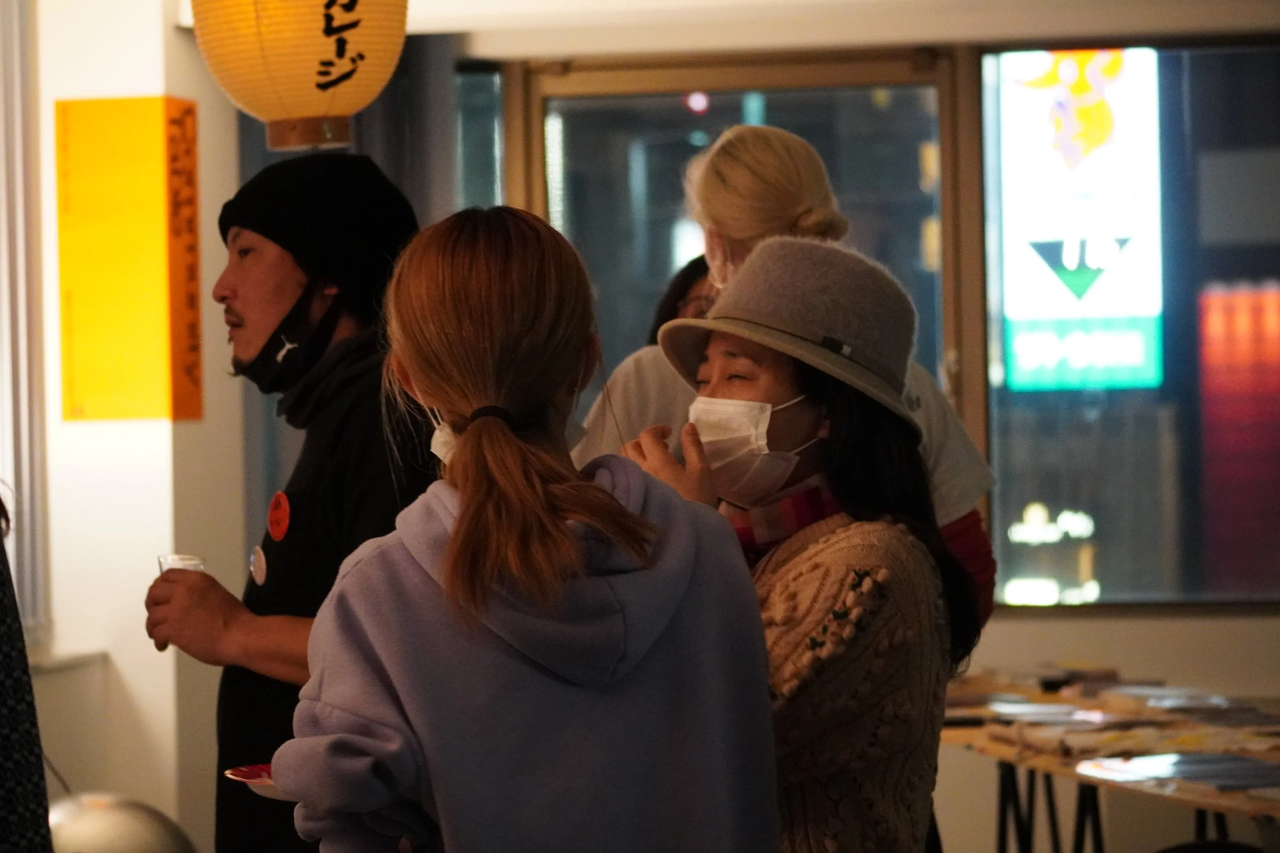 室内で会話をしている複数の人々。女性はマスクを着用し、帽子をかぶっている。背景には窓といくつかの掲示板や看板が見える。