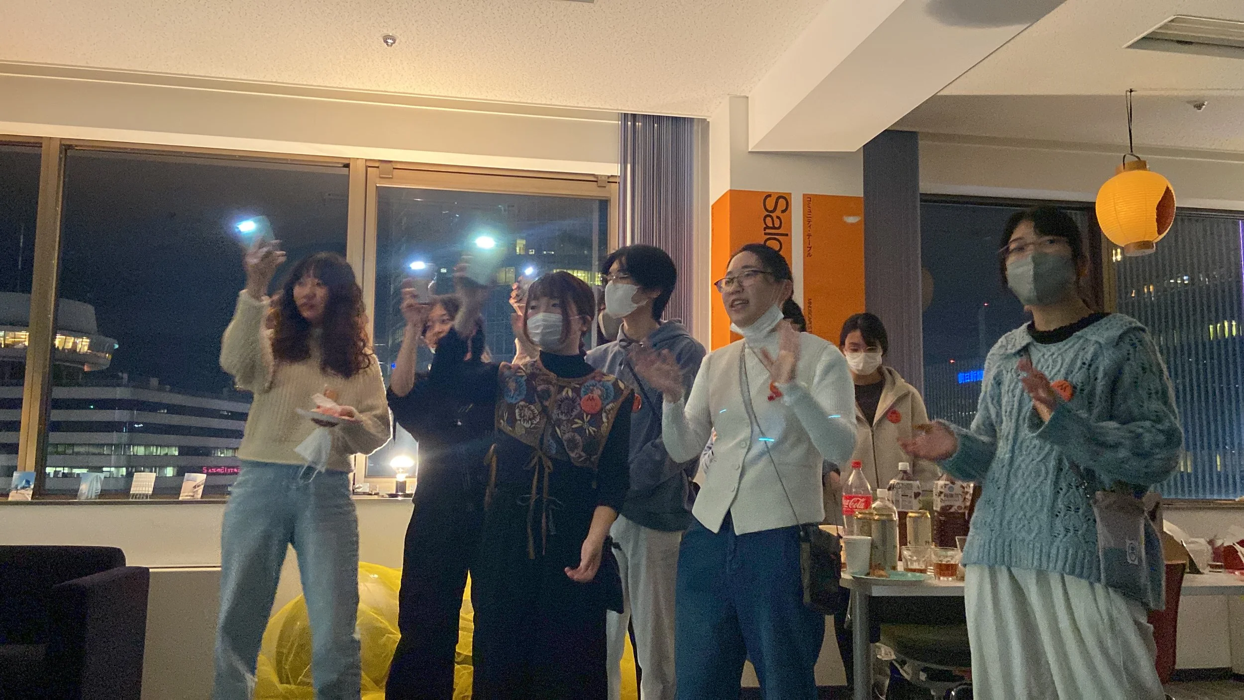夜の室内で、マスクをした女性たちが集まり、お祝いをしている様子。