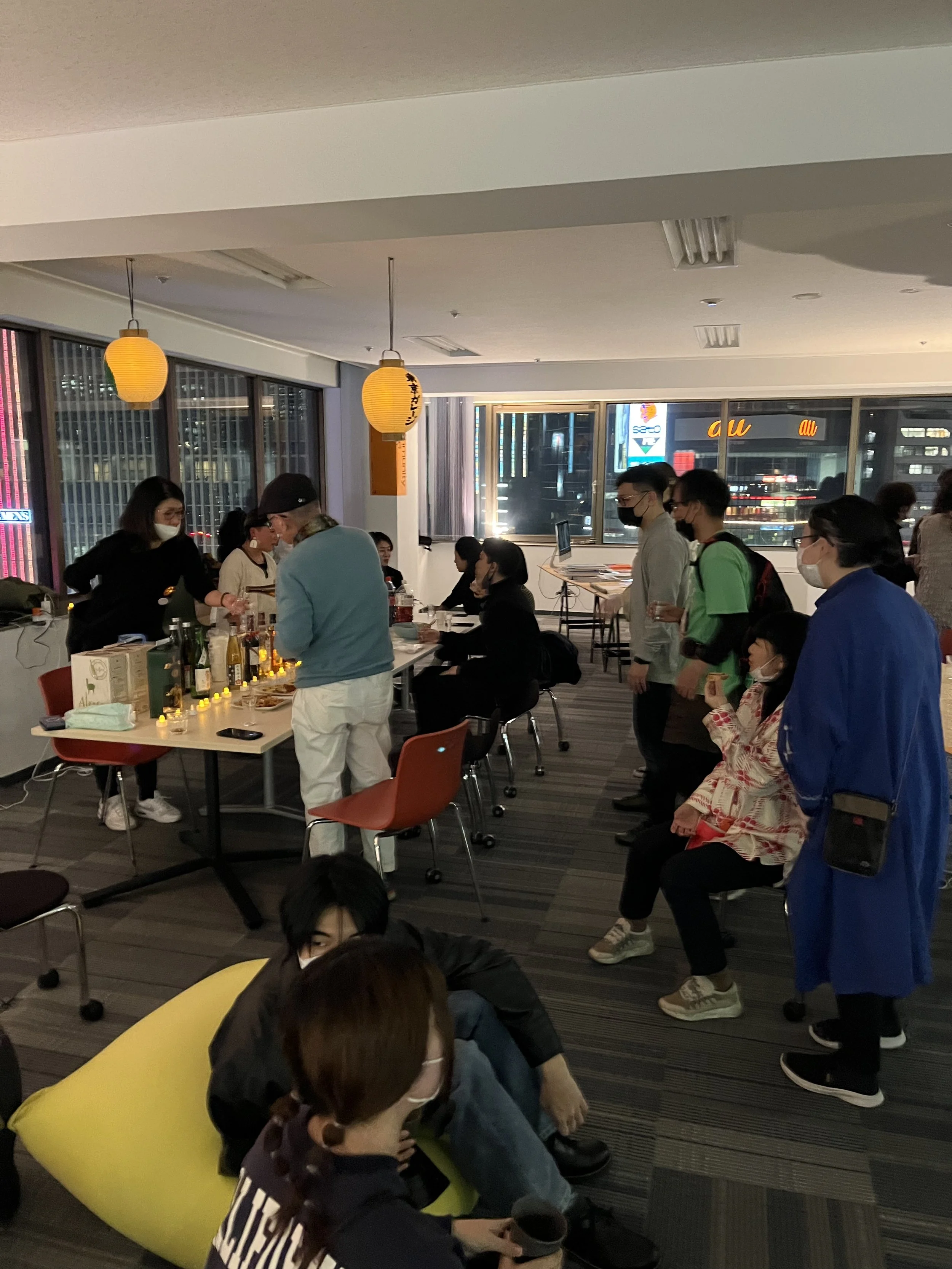 室内のパーティーやイベントの様子で、テーブルに飲み物や食べ物、キャンドルが並び、参加者が並んだり座ったりしている。窓の外にはネオン看板や街の景色が見える。