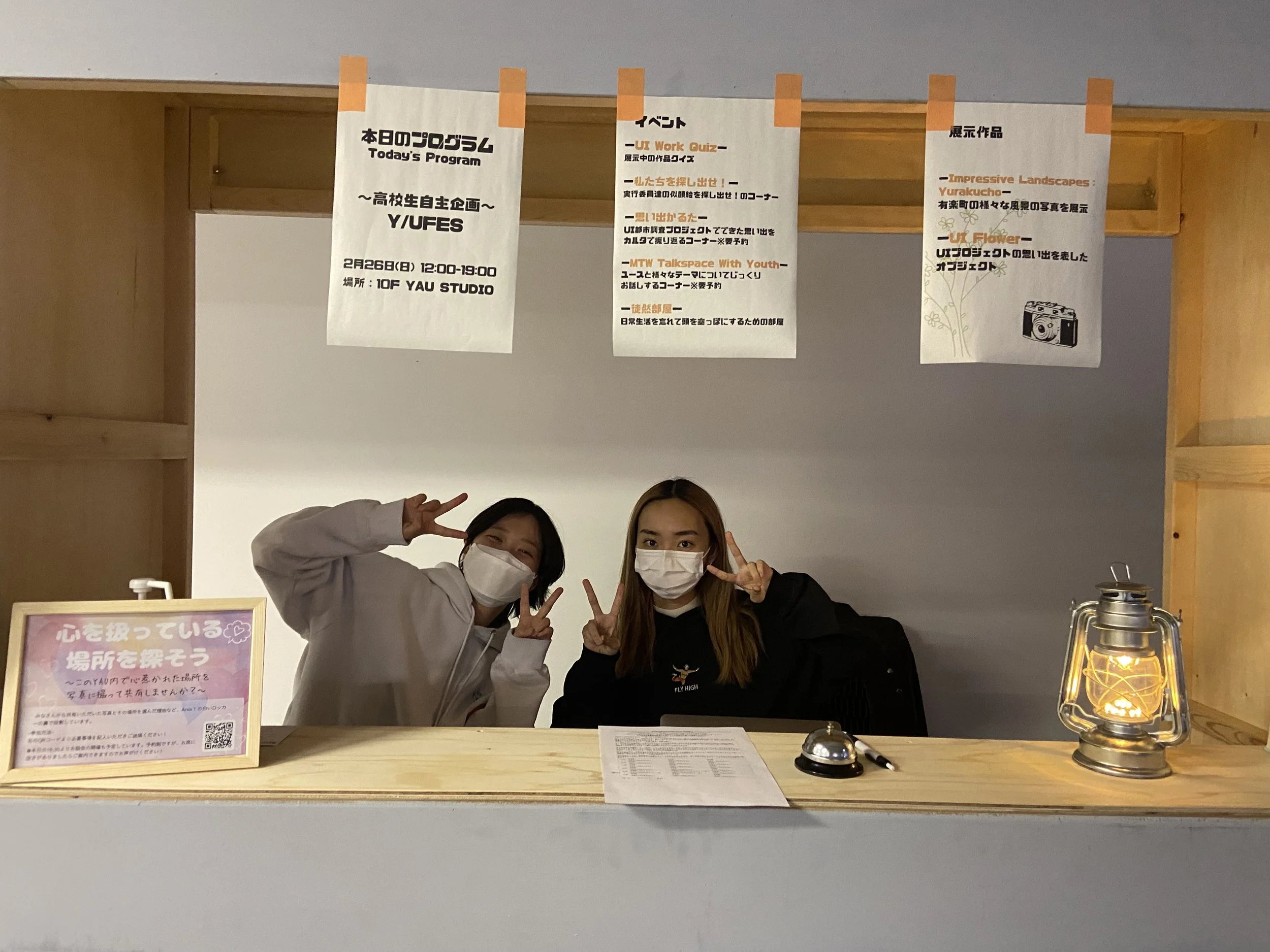 受付のカウンターに二人の女性が座っていて、二人ともマスクをしている。背景には掲示板に日程やイベント内容の掲示が貼ってあり、左側には心を癒している場所をテーマにした案内板とキャンドルスタイルのランプが置かれている。