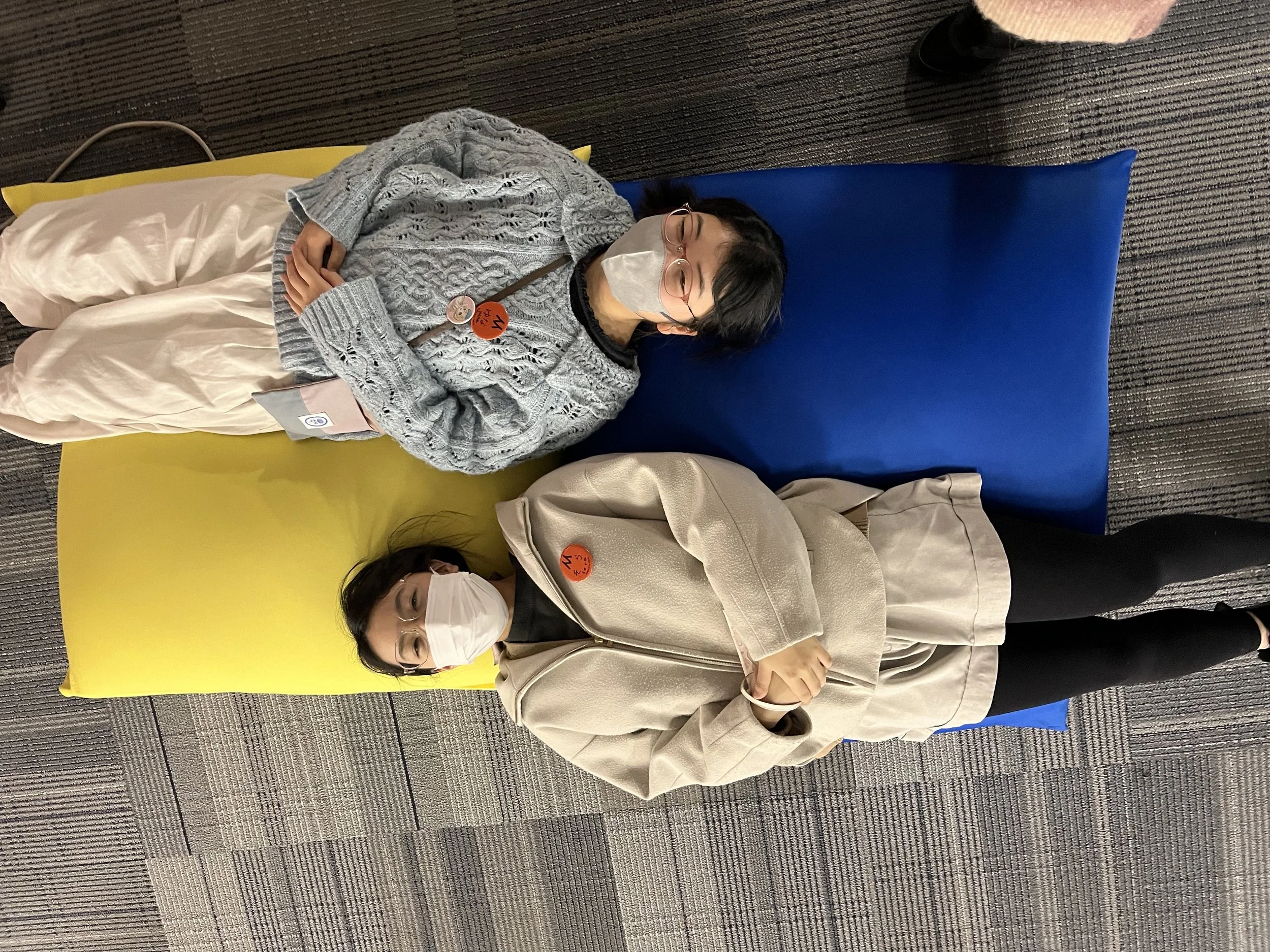 黄色と青の背もたれのパッドに横たわる二人の女性。二人ともマスクを着用しメガネをかけている。
