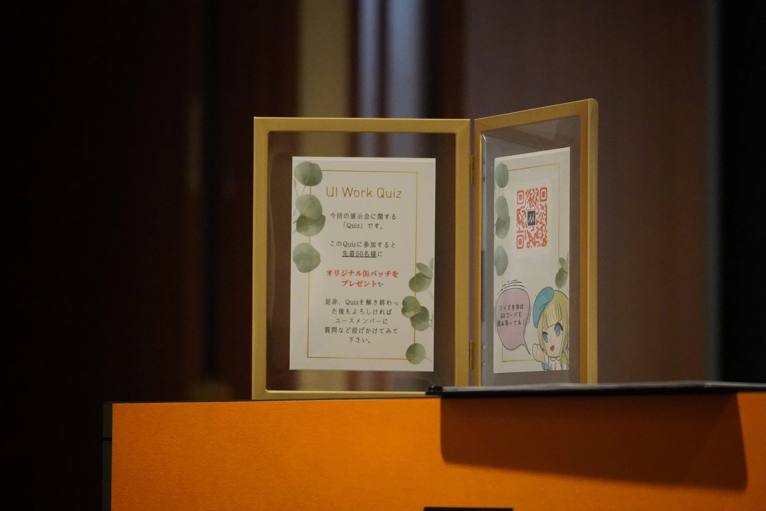 オレンジ色の展示台の上に設置された二つのスタンドを囲む透明なケースの中に、紙の掲示物とQRコードが表示されている。左側には「UI Work Quiz」と書かれた案内と葉の模様が描かれている。