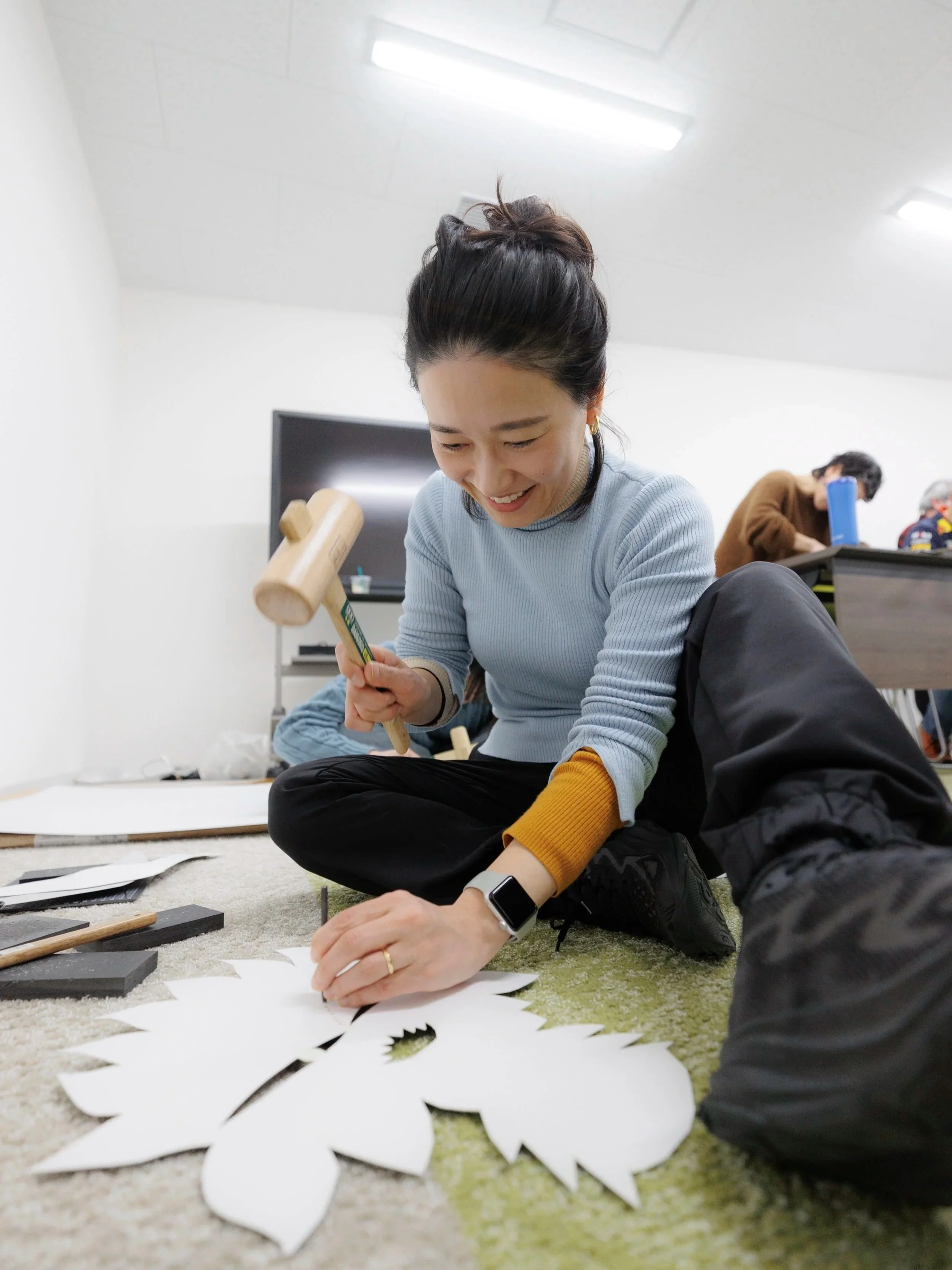 床に座って紙に絵を描いている笑顔の女性と、背景には男性と子供がいる室内の風景