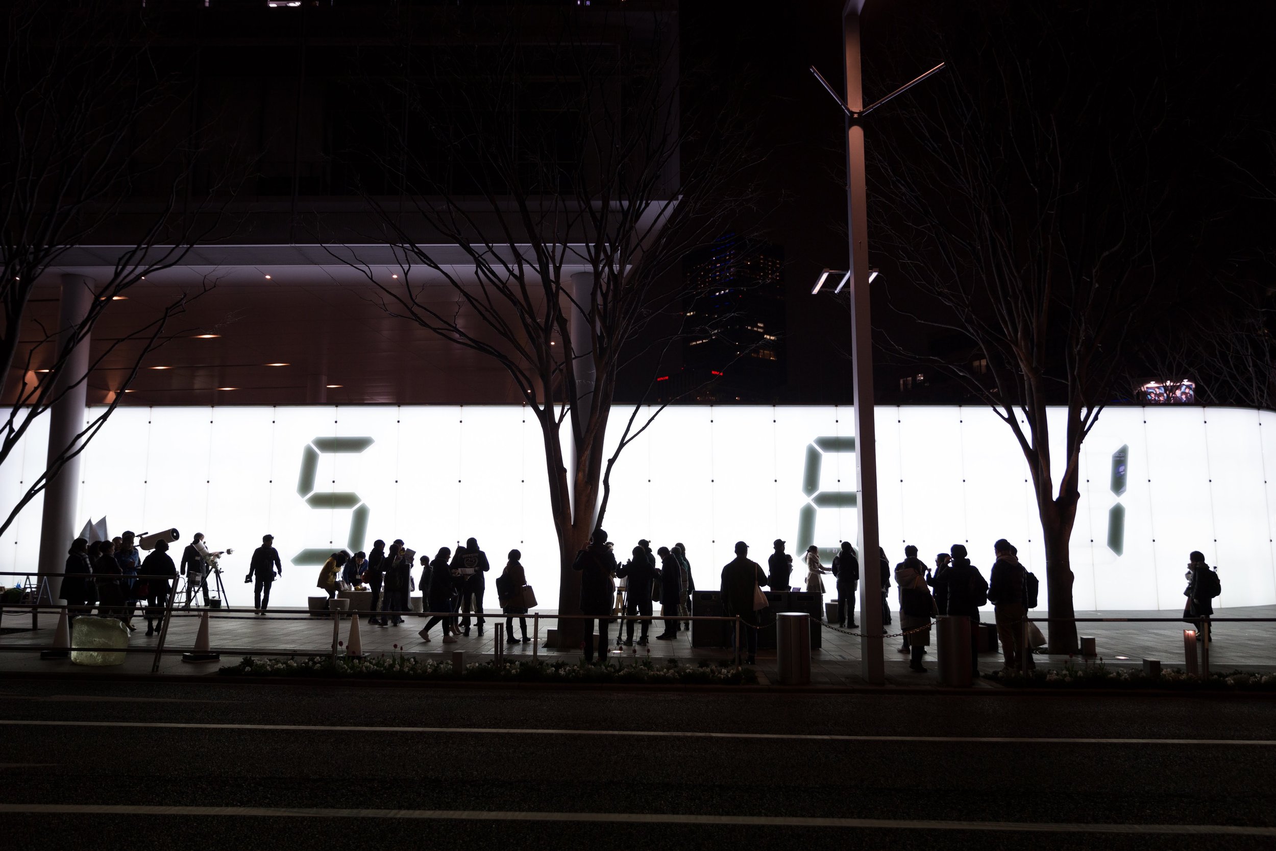 夜の六本木のテレビ朝日前を人々が行き交う光景。背後には宮島達男の代表的な作品「Counter Void」の数字が大きく映し出されている。