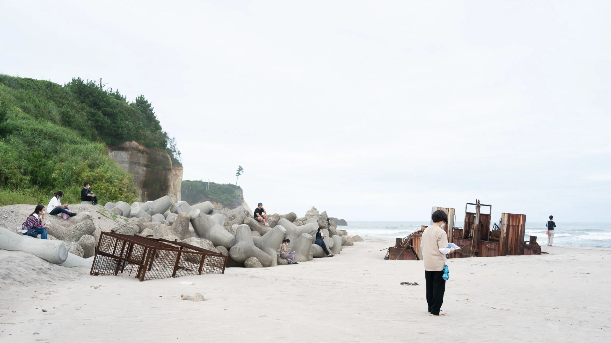 海岸で人々が座ったり歩いたりしている風景。岩や海、津波で座礁して放置された古い船の残骸が見える。背景には緑の丘と空が広がる.