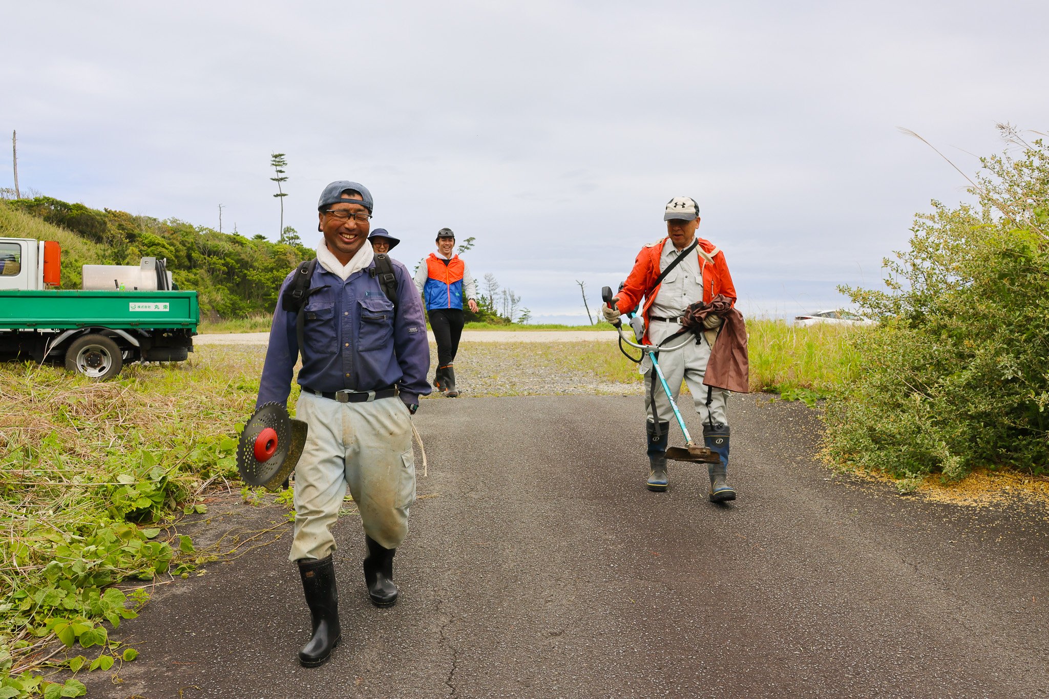 緑豊かな自然の中で、大人の男女4人がボランティア活動を楽しんでいる。彼らは笑顔で、道具や作業用品を持ち、カジュアルなアウトドア服装をしている。