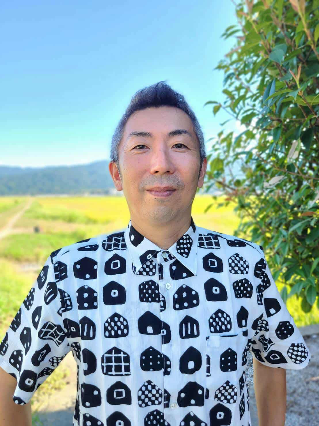 自然の中で笑顔の男性のアップ写真。青空と緑の木々、田畑が背景に広がる風景。男性は白黒の幾何学模様のシャツを着ている。