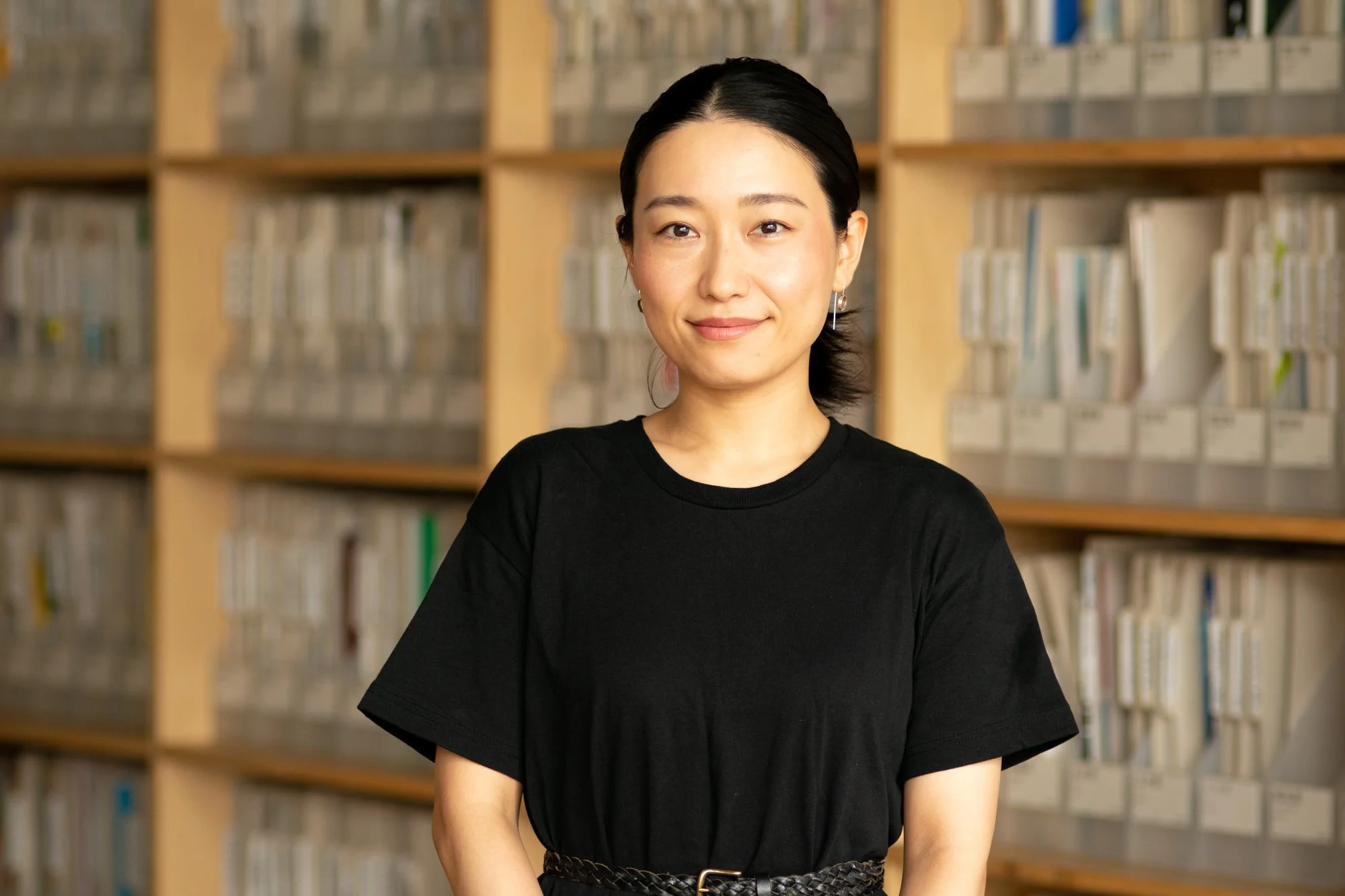 書棚の前で微笑む若い女性。短い黒髪で黒いTシャツを着ている。