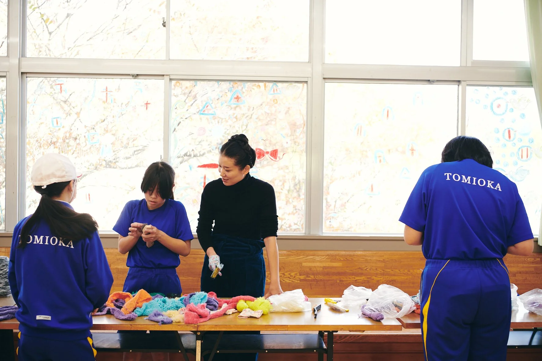 教室でハンドメイドの編み物をしている学生と先生。学生は青い制服を着ており、先生は黒い服を着ている。窓の外には木々の葉や装飾が見える。