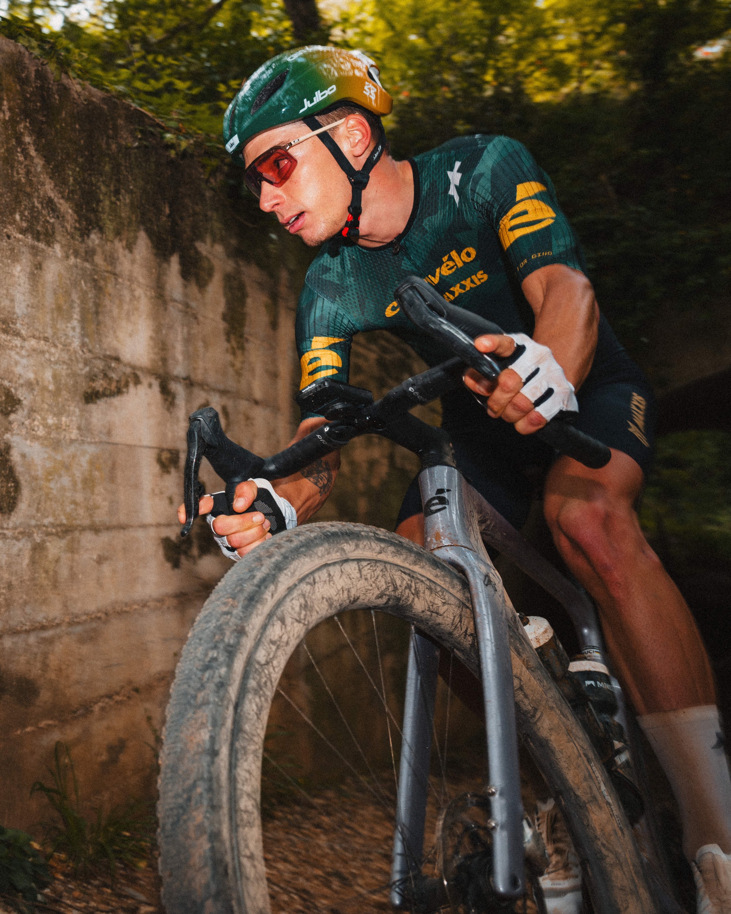 A male mountain biker wearing a green helmet, red sunglasses, and a teal cycling jersey is riding a dirt trail through a wooded area. He is focused on steering his bike around a curve with a rough, muddy tire.