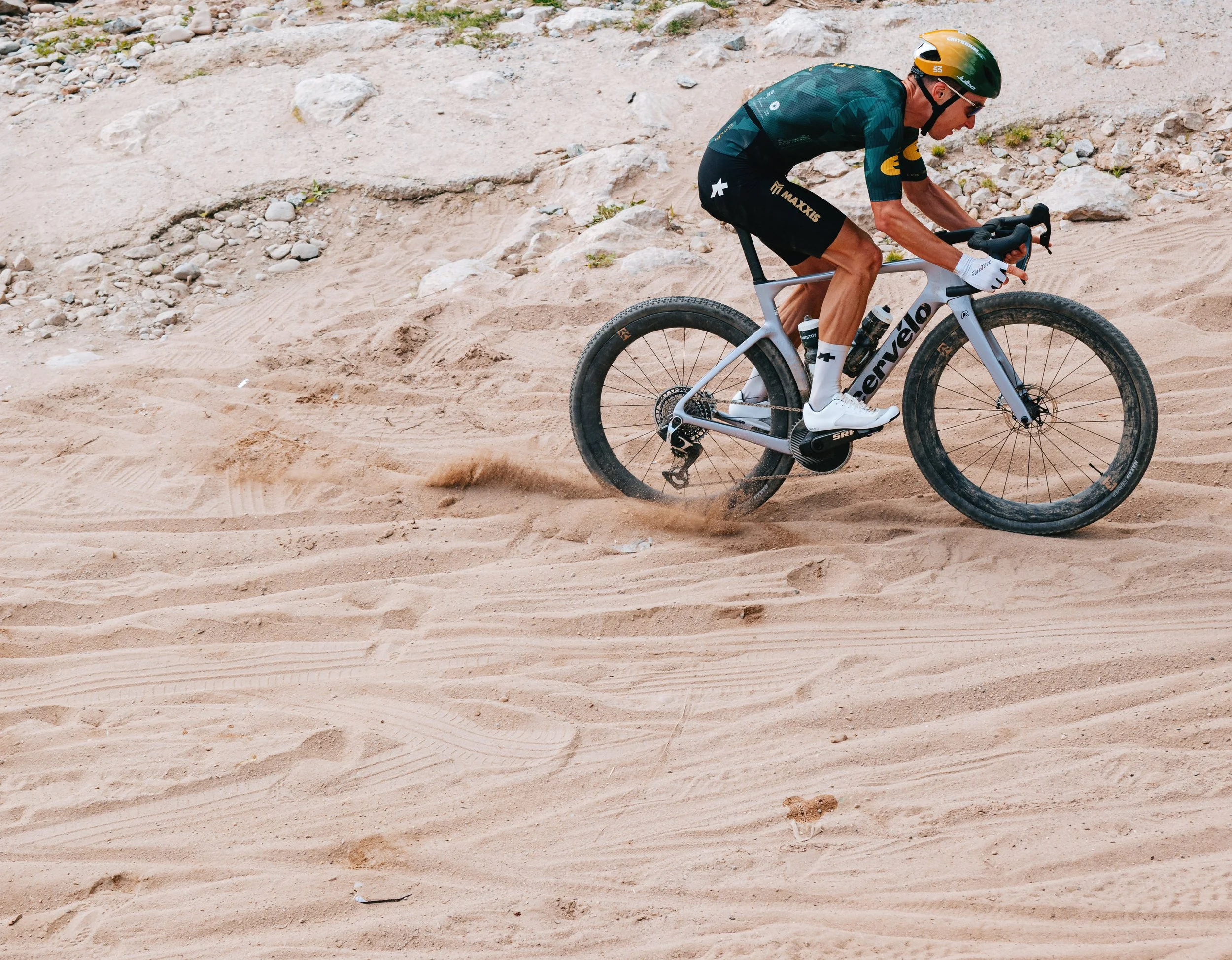 A person riding a gravel bike on a sandy, rocky trail. The rider is wearing a helmet, sunglasses, cycling gloves, and athletic clothing, leaning forward while pedaling.