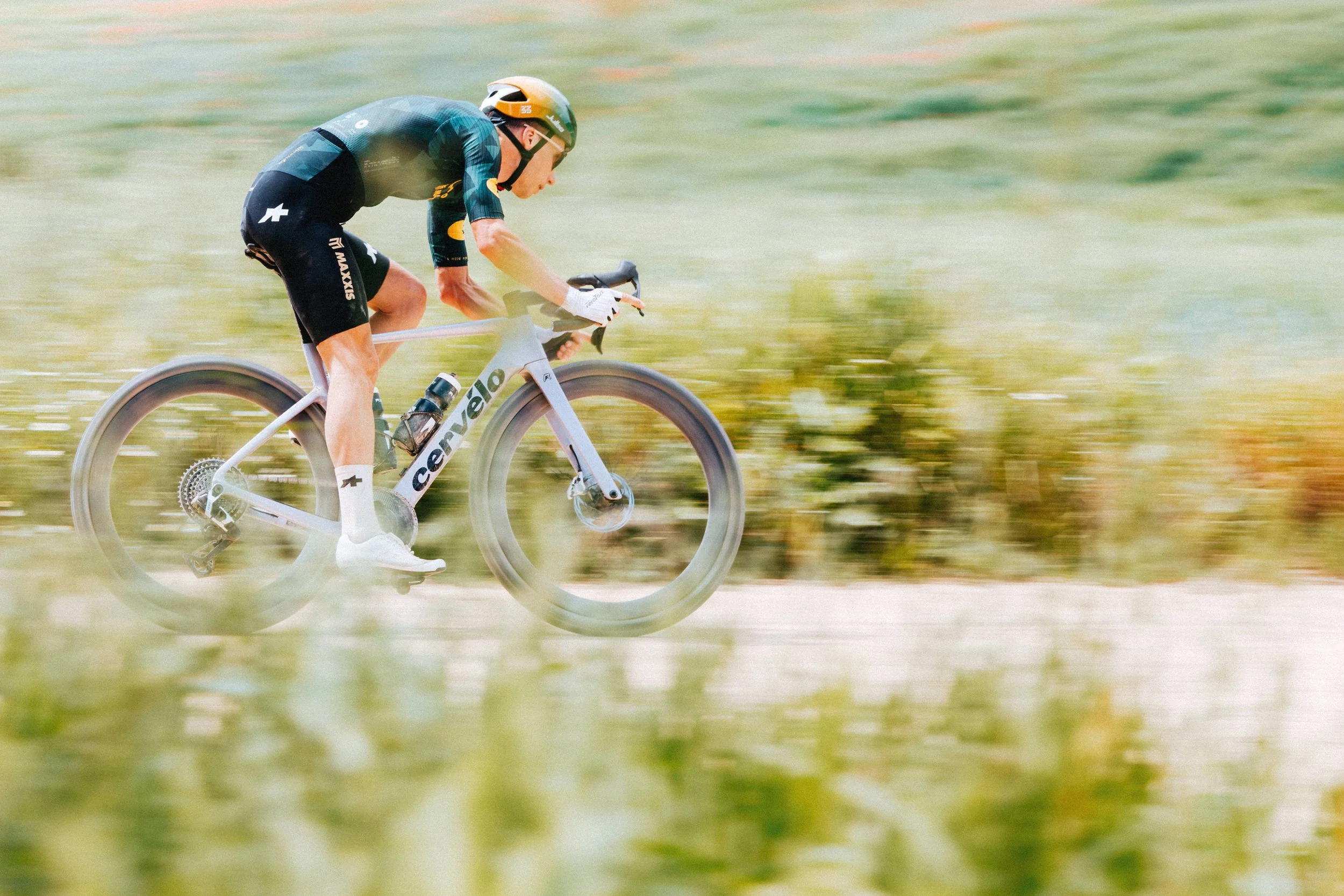 A cyclist riding a white bicycle on a grassy outdoor trail, wearing a black helmet, black and blue racing outfit, and white gloves.