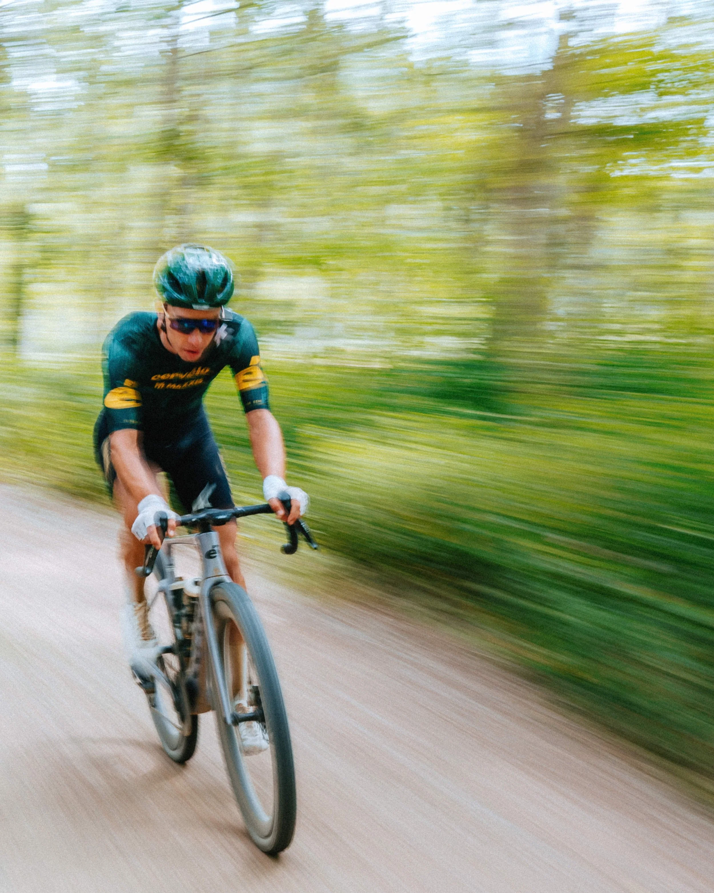 A cyclist wearing a helmet, sunglasses, and a dark cycling jersey riding a bicycle on a wooded trail with motion blur indicating speed.