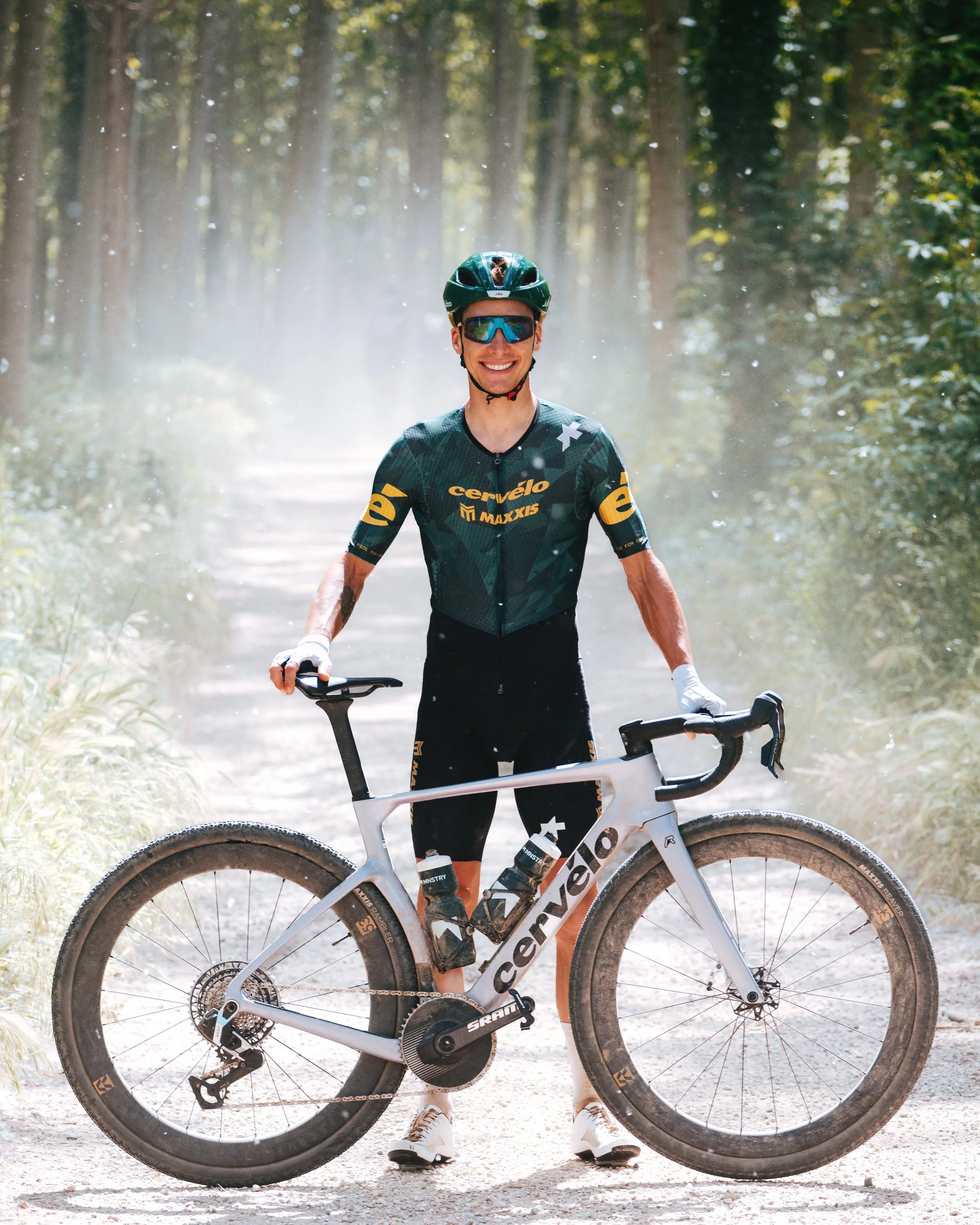 A smiling man standing outdoors in a forest holding a mountain bike, wearing a green cycling helmet, sunglasses, a jersey, cycling shorts, and shoes.