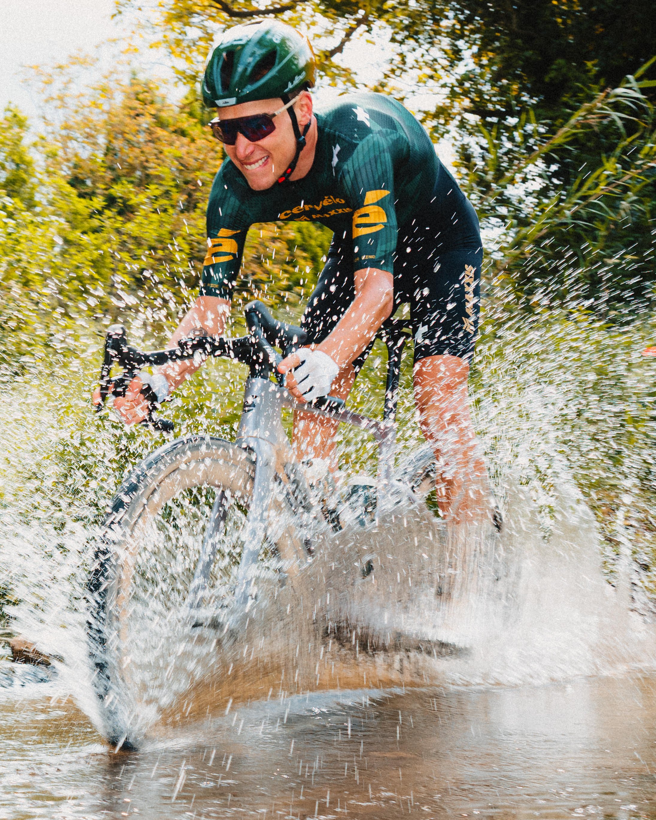 A man riding a bicycle through water splashing around, outdoors with trees and foliage in the background.