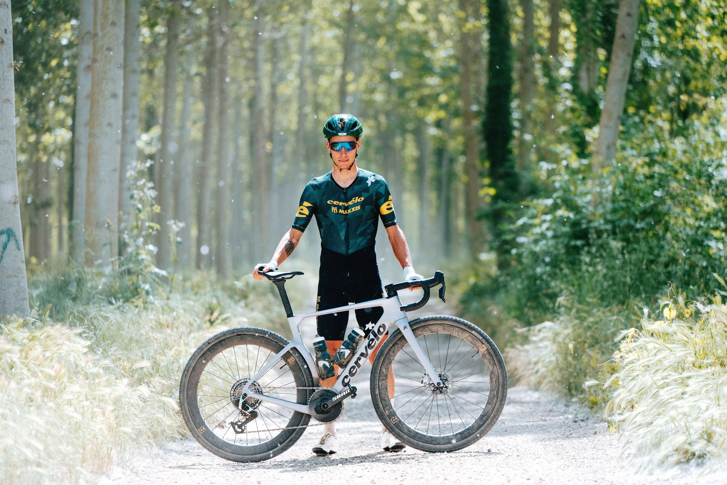 A male cyclist wearing a helmet, sunglasses, a cycling jersey, and shorts stands outdoors in a forest with a mountain bike, with trees and sunlight in the background.