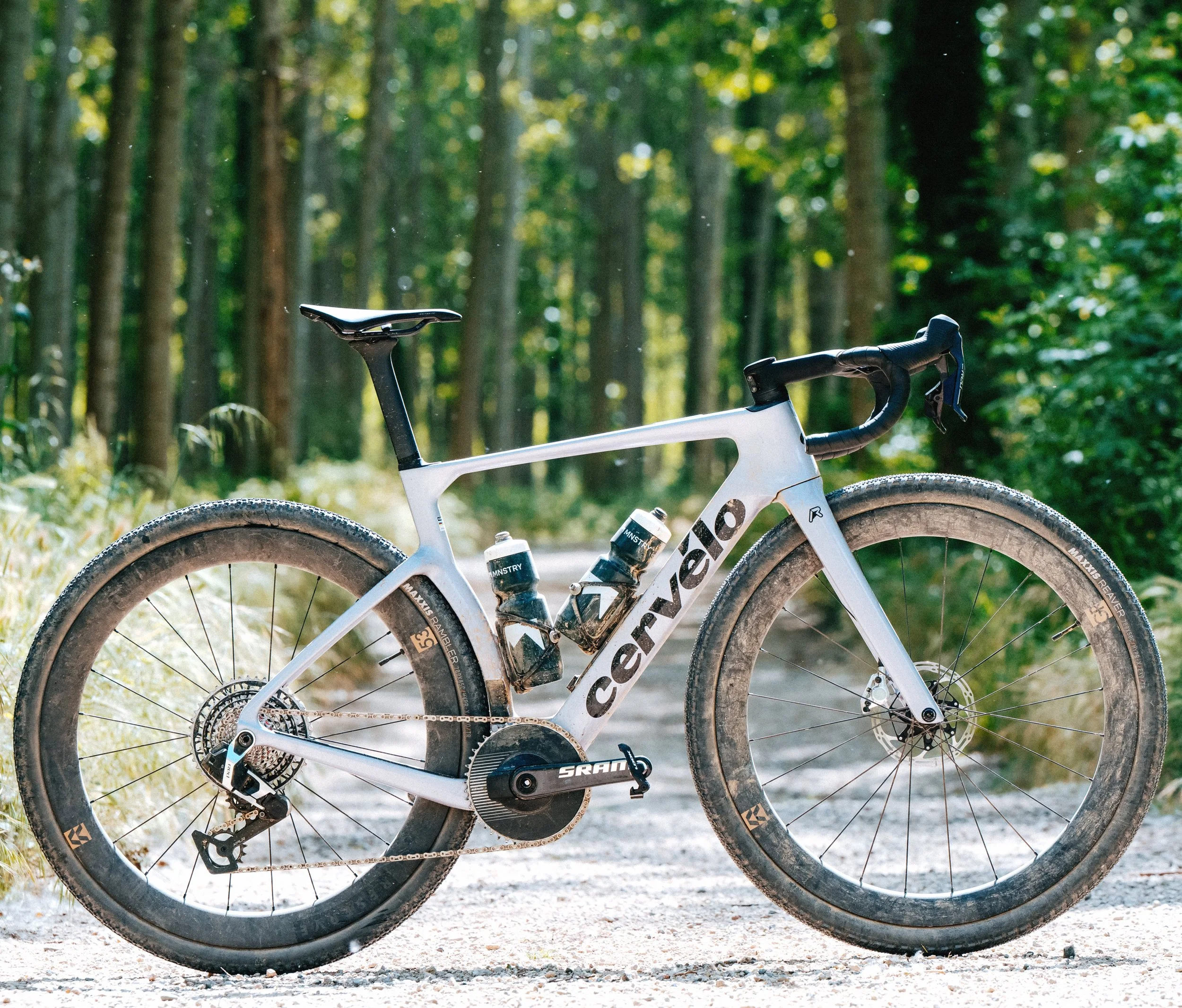 Close-up of a white Cannondale mountain bike with black accents, muddy tires, on a dirt trail among a green forest.