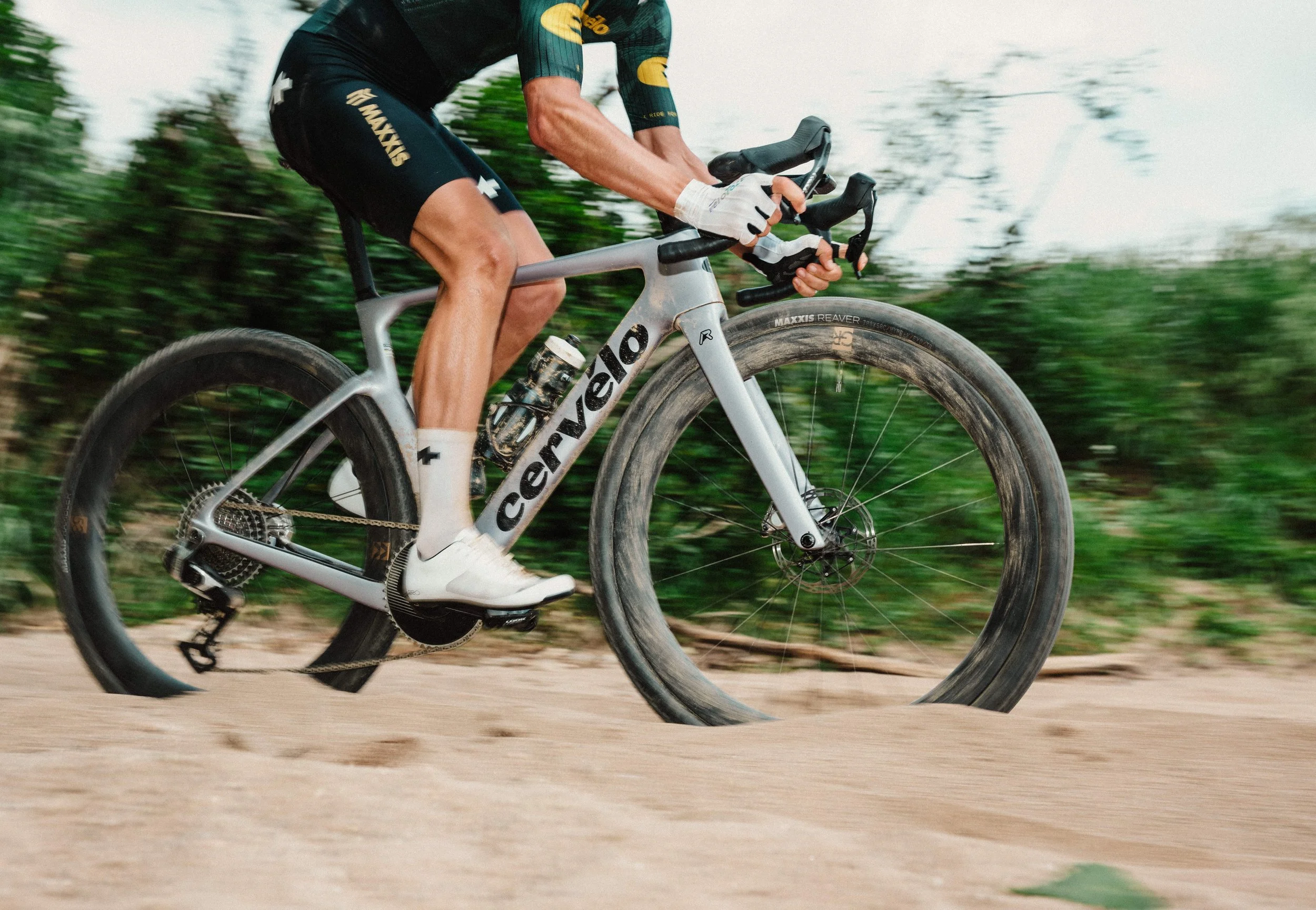 A cyclist riding on a dirt trail with a blurred green background, wearing a black and yellow cycling jersey, black shorts, and white shoes.
