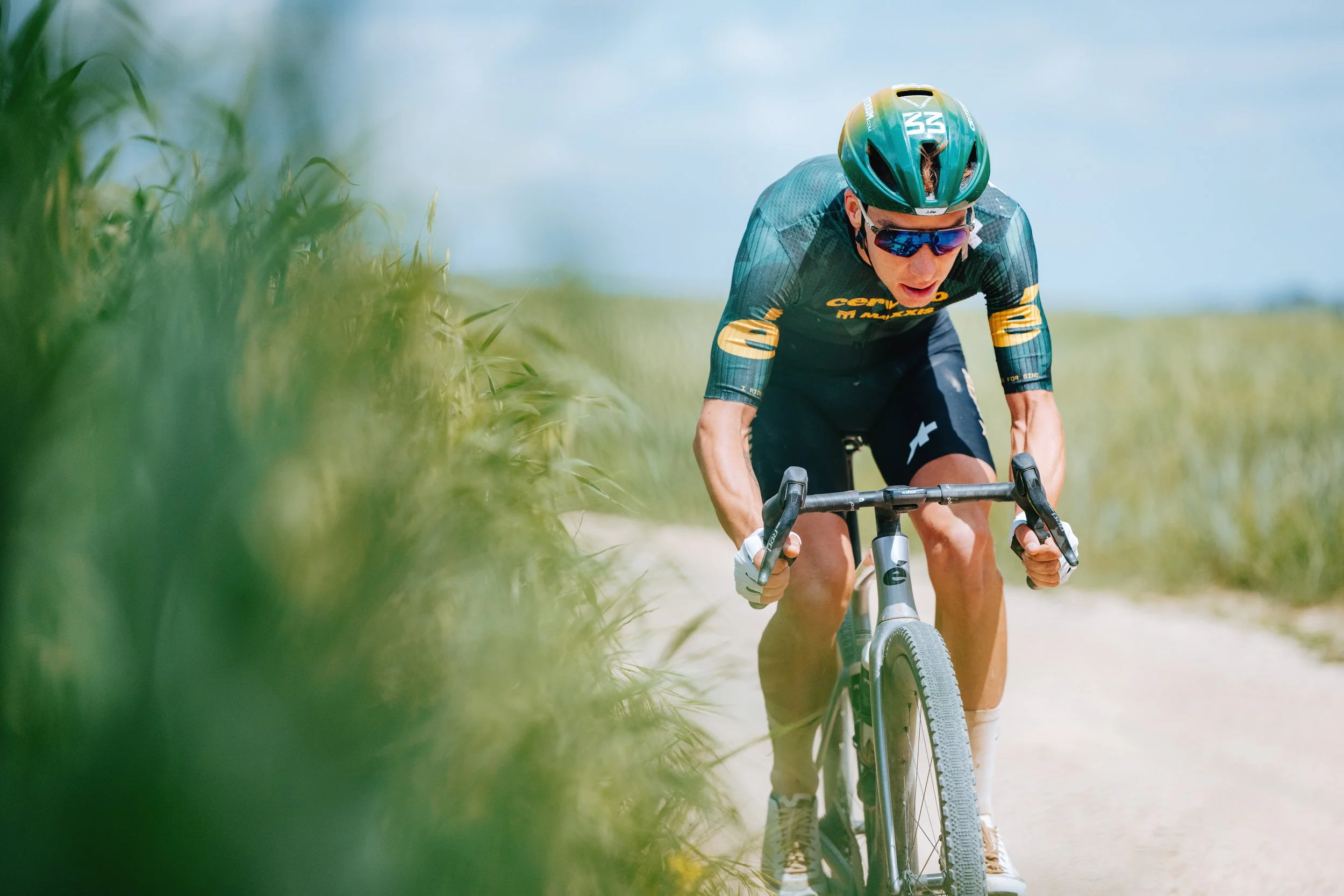 A male cyclist in sunglasses and a helmet riding a mountain bike on a dirt trail through a grassy area under a blue sky.