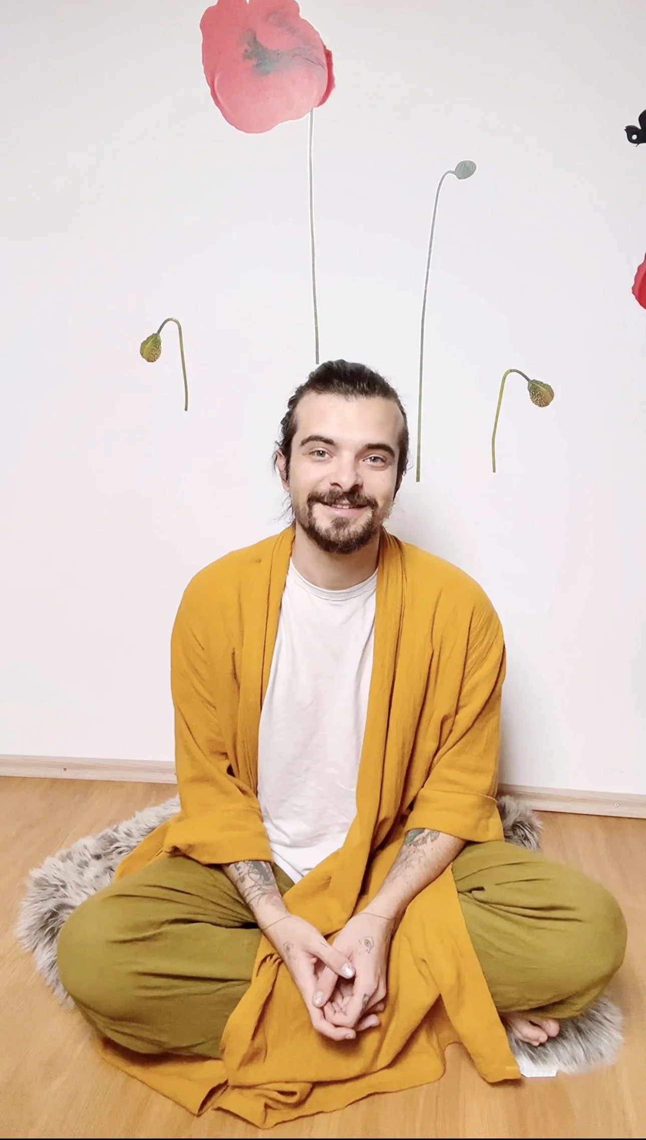 A young man with a beard and tattoos on his hands, wearing a white t-shirt and a mustard yellow jacket and pants, sitting cross-legged on a furry rug on a wooden floor, smiling at the camera. Behind him on a white wall are decorative paper flowers and butterflies.