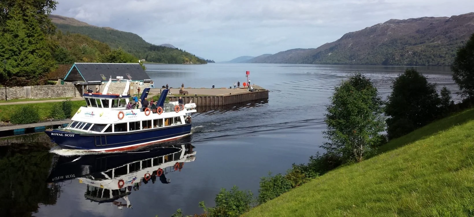 A boat named Royal Scot sailing on a calm lake with hills and mountains in the background, showing a dock with people and a small building nearby.