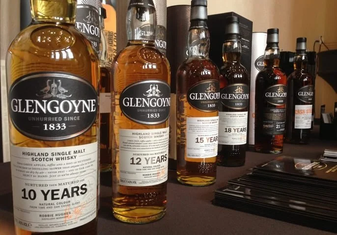 A lineup of GlenGoyne single malt Scotch whiskey bottles on display, with various age statements including 10, 12, 15, 18, 21, and 25 years, on a table with black pamphlets or brochures.